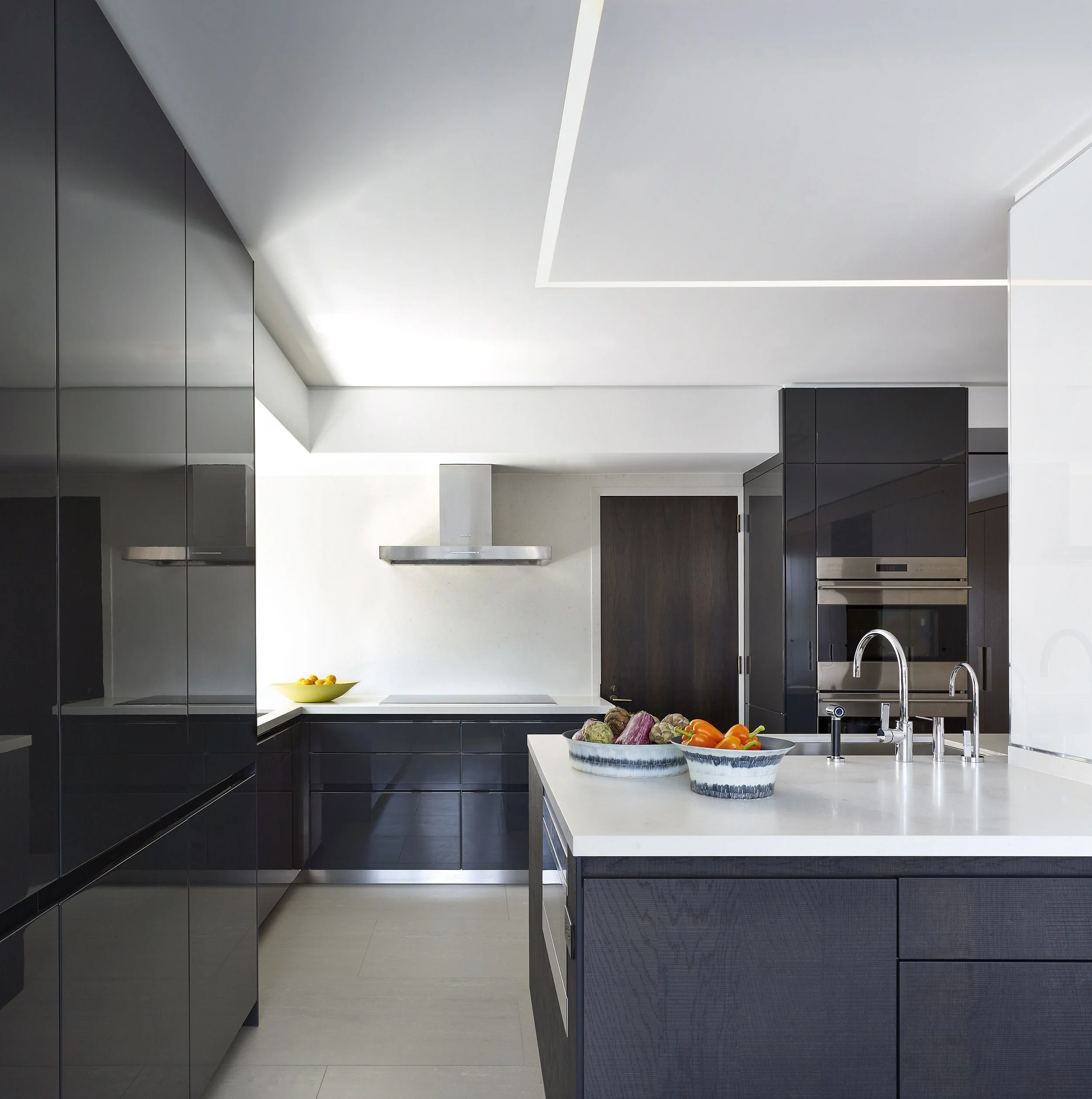Modern kitchen with black cabinetry, white countertops, and stainless steel appliances. Fruits and vegetables in bowls on the island and counter. Sleek, minimalist design with clean lines and neutral colors.