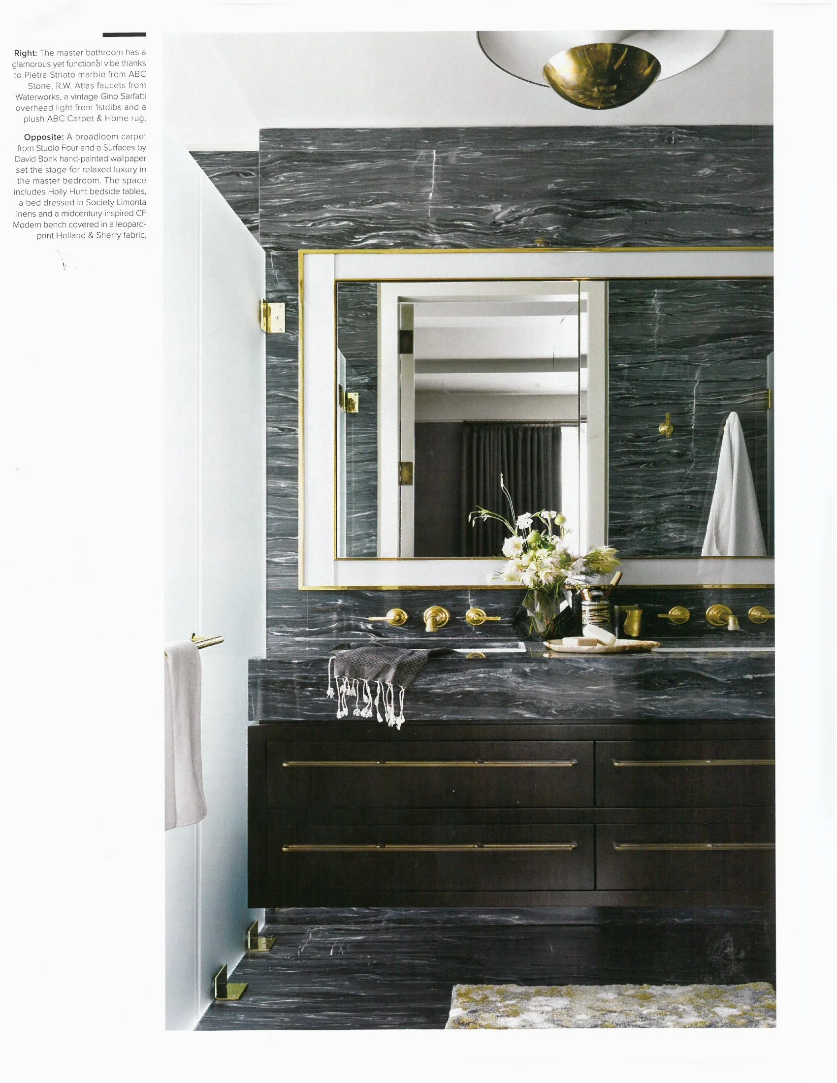 A luxurious bathroom featuring bold black marble walls and countertop, complemented by gold fixtures and a large mirror. The vanity has dark wood drawers with gold handles, and decor includes a vase of flowers and neatly arranged toiletries.