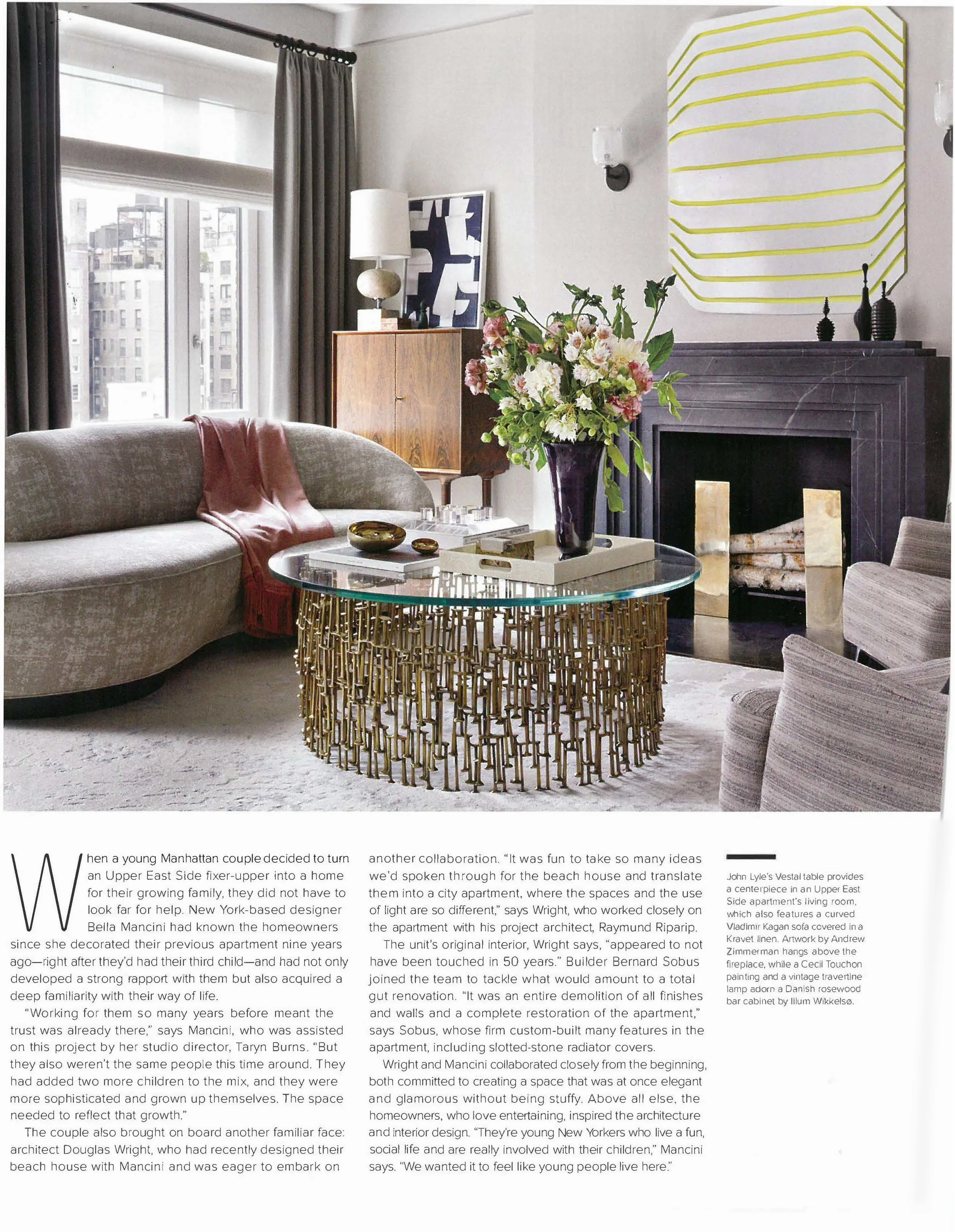 A living room with a grey curved sofa, a glass coffee table with a gold wire base, a vase of flowers, and various decorative items. There are large windows with grey curtains, a wooden cabinet, a framed artwork, a fireplace, and modern wall sconces.