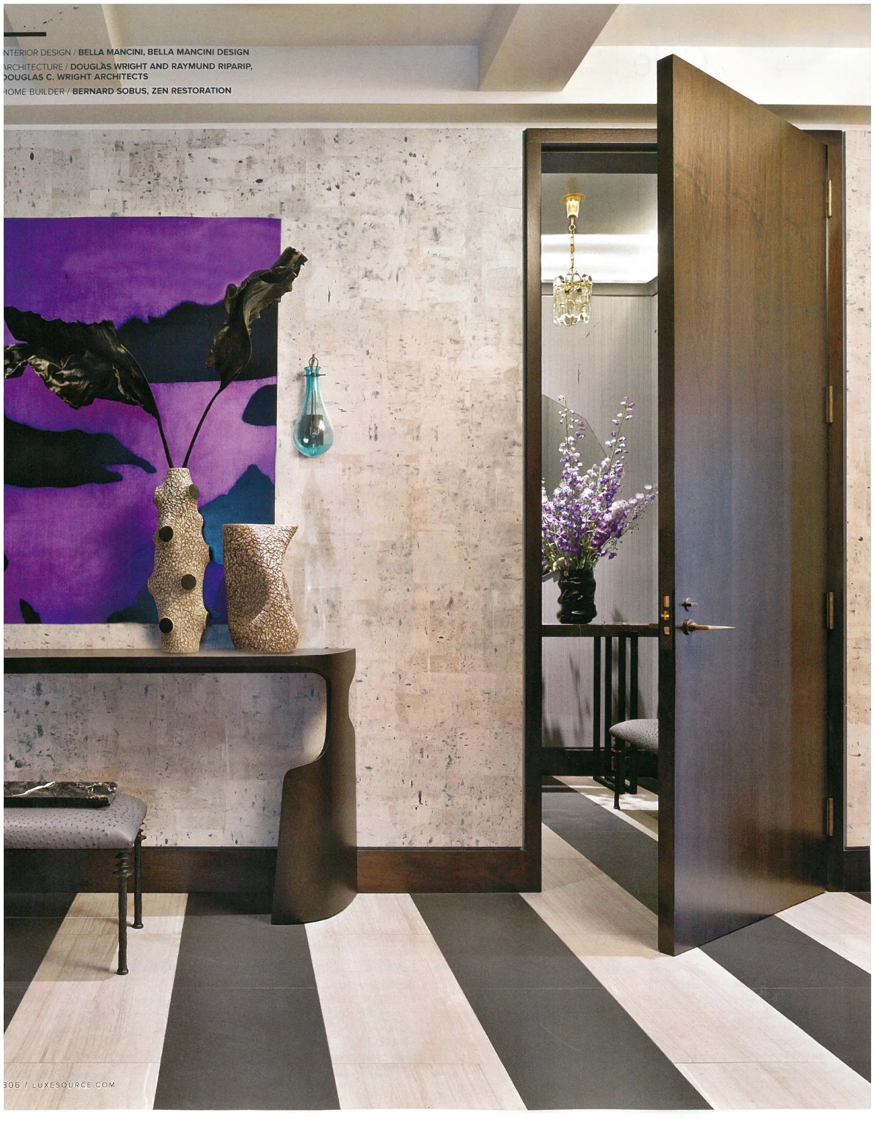 Interior room with a textured white wall, a dark wooden side table with vases, a colorful abstract art piece, an open wooden door leading to a room with a chandelier and purple flowers in a black vase, and a black and white striped floor.