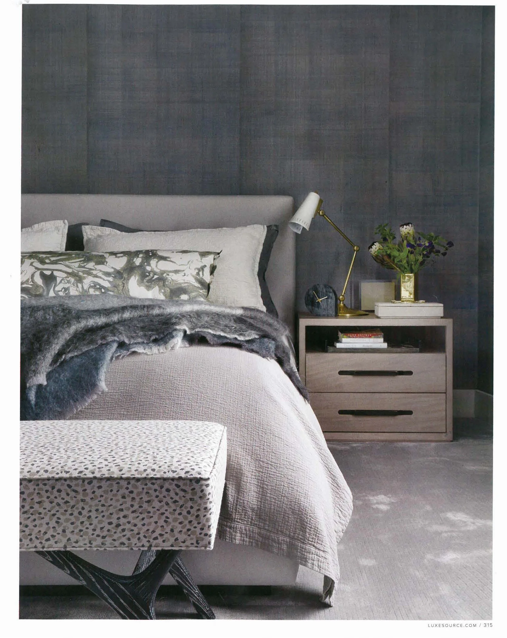 A modern bedroom featuring a bed with white linens, a gray upholstered headboard, and a faux fur throw. A small nightstand with a white finish holds a gold desk lamp, a vase with flowers, and a stack of books. The wall behind the bed is covered in da