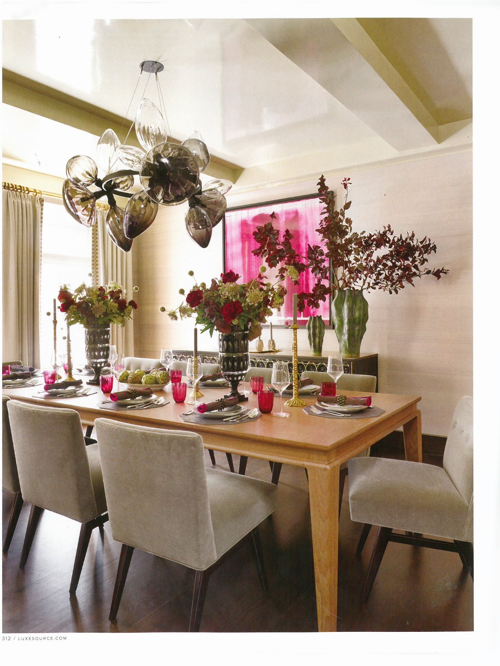 A dining room with a wooden table set with plates, glasses, and pink cups. Vases with large floral arrangements and candles are on the table. A modern chandelier hangs from the ceiling. Light-colored chairs surround the table, and cream-colored curta