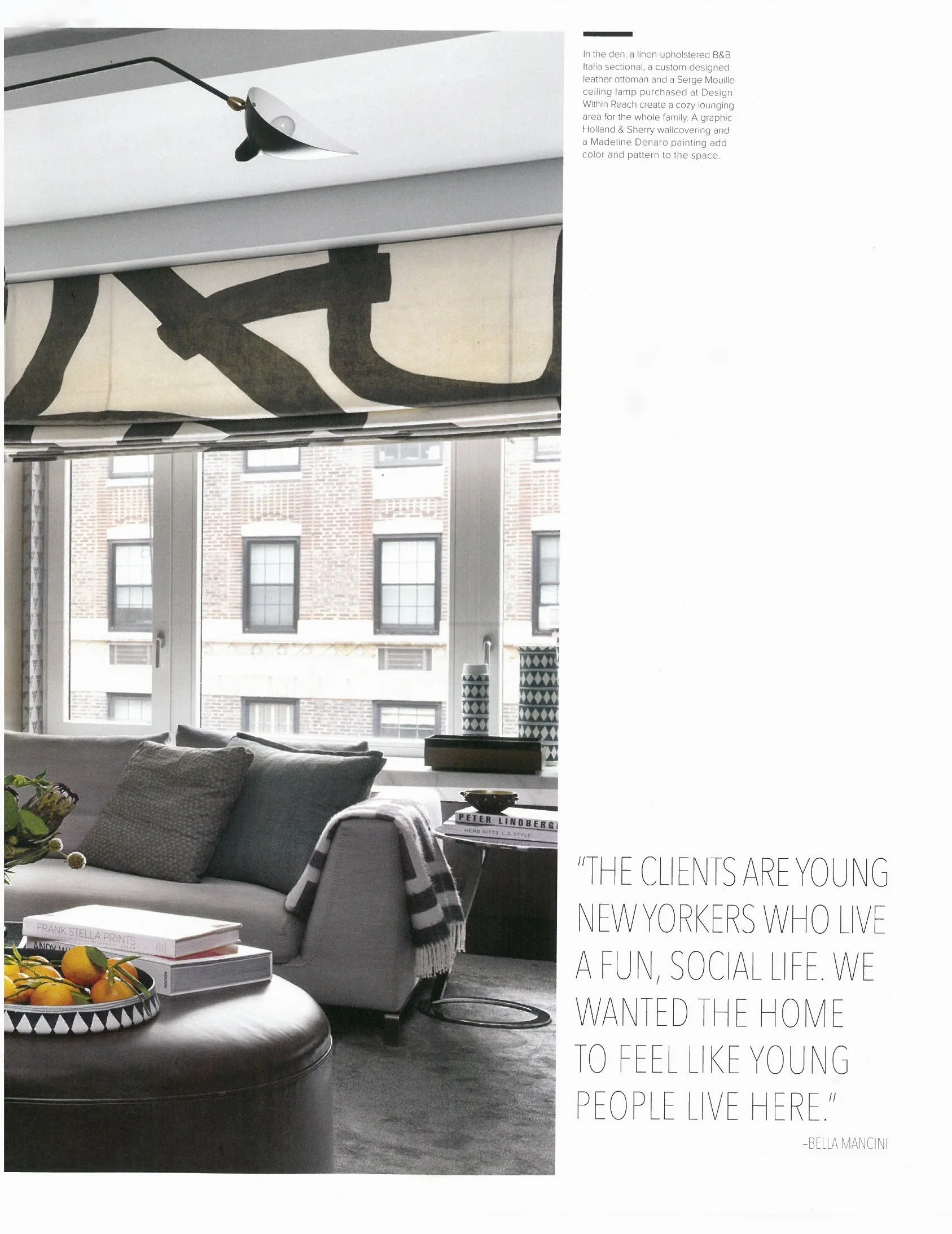 Living room with gray sofa, black and white striped throw blanket, coffee table with books and a bowl of oranges, large window with black and white patterned curtains, decorative vases, and city view.