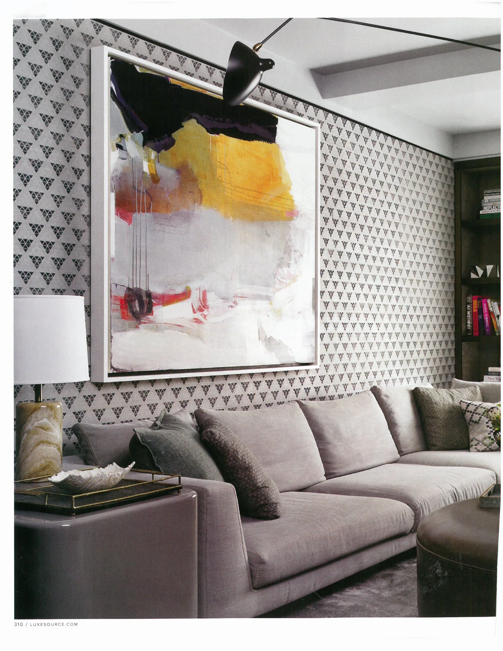Living room with patterned wallpaper, a large abstract painting on the wall, a gray sofa with multiple textured pillows, a side table with a lamp and decorative bowl, and a black bookshelf in the background.