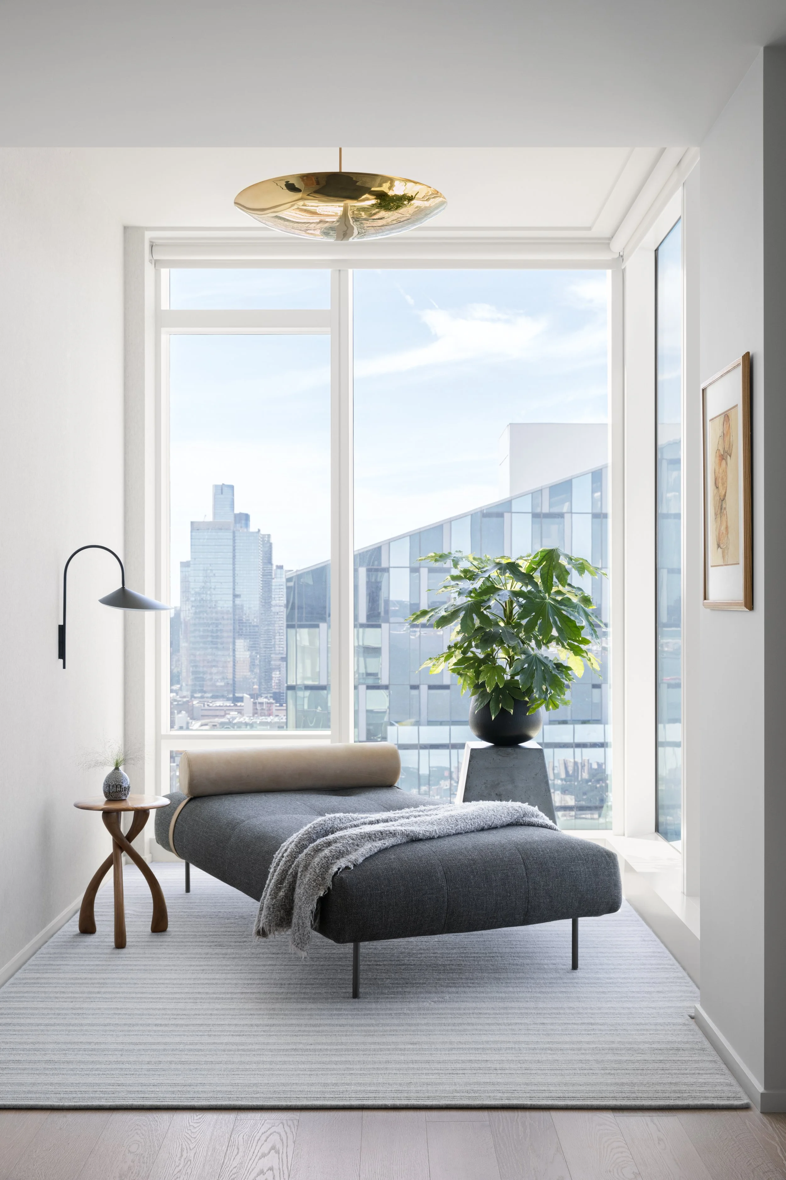 Modern interior room with a large window, a gray chaise lounge, a small side table with a decorative item, a large potted plant, a wall-mounted lamp, and artwork on the wall, with city buildings visible through the window.