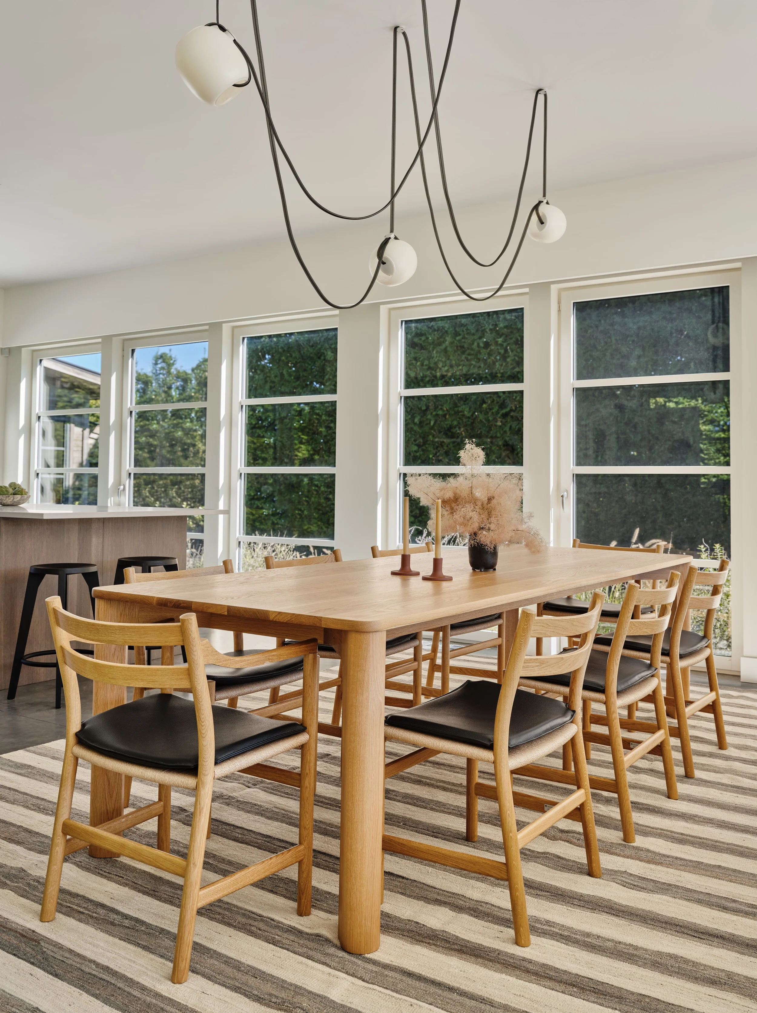 A modern dining room with a large wooden table surrounded by eight matching wooden chairs with black cushioned seats. The room has large windows allowing natural light and showing greenery outside. A black candle holder with two yellow candles and a 