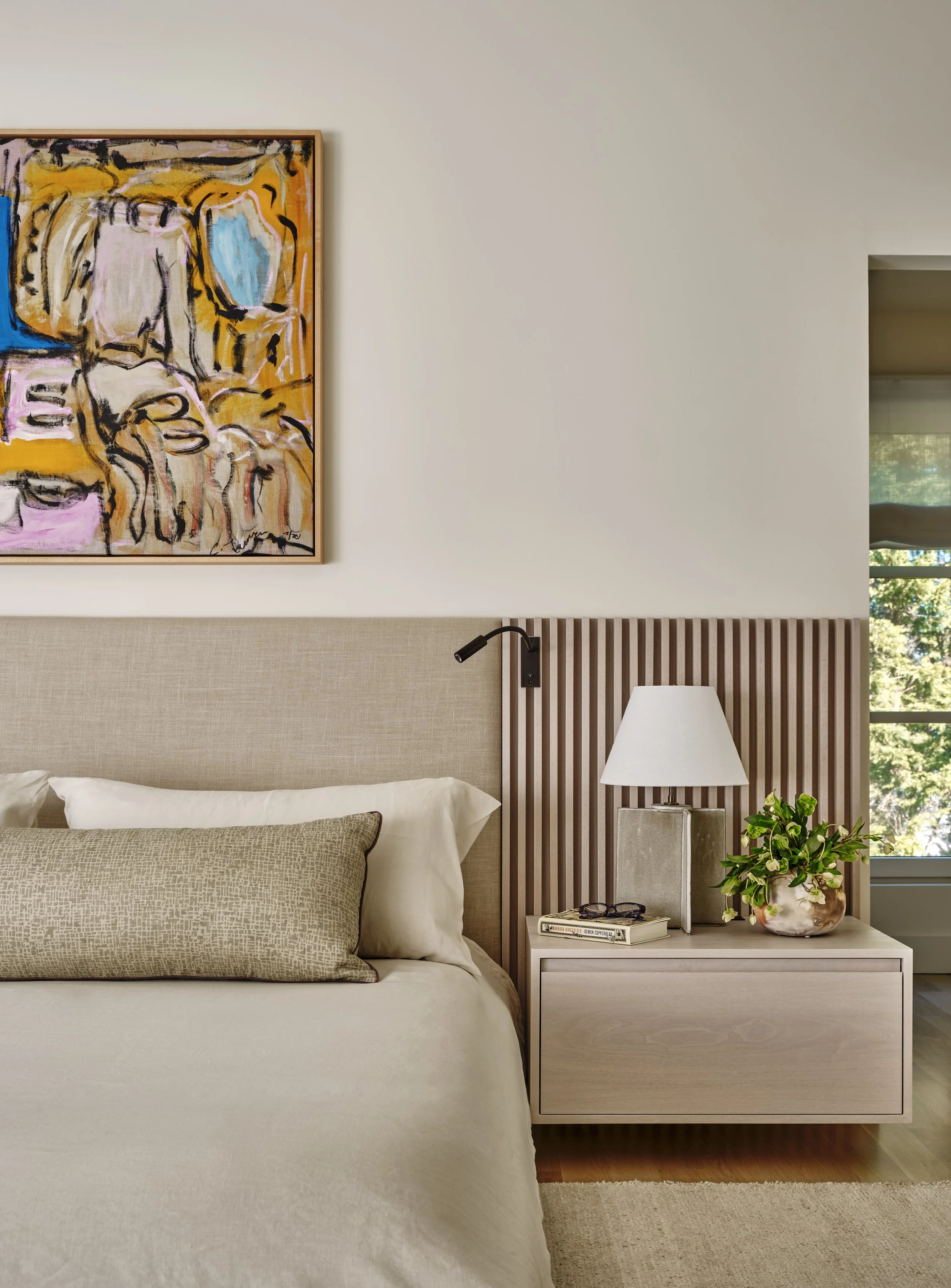 A bedroom with a beige headboard, a beige pillow, and a white pillow. There is a nightstand with a lamp, a potted plant, and magazines. An abstract painting hangs above the bed, and there is a window showing greenery outside.