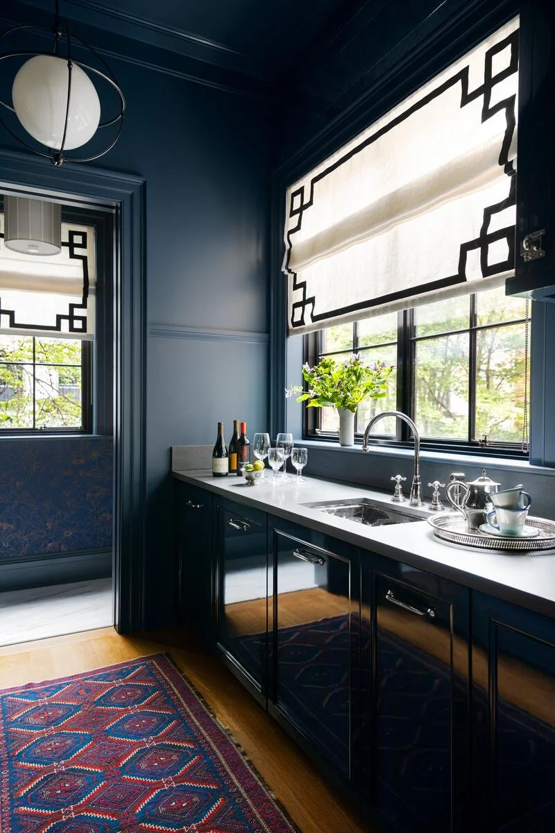 A kitchen with dark blue cabinetry and walls, a white countertop, a window with black geometric patterned blinds, a vase with green flowers, wine bottles, glasses, and a tray with teapots and cups. A patterned rug on the wooden floor.