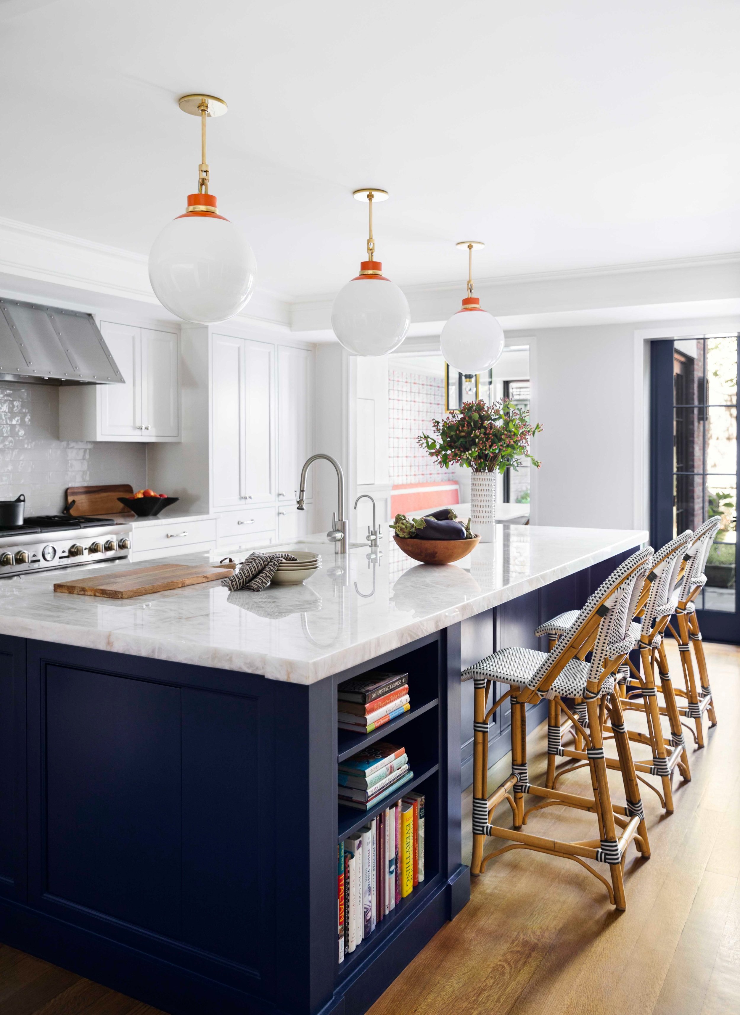 Modern kitchen with white cabinets, navy blue island with open book shelves, white marble countertop, three hanging pendant lights, wicker chairs, and a sliding glass door.