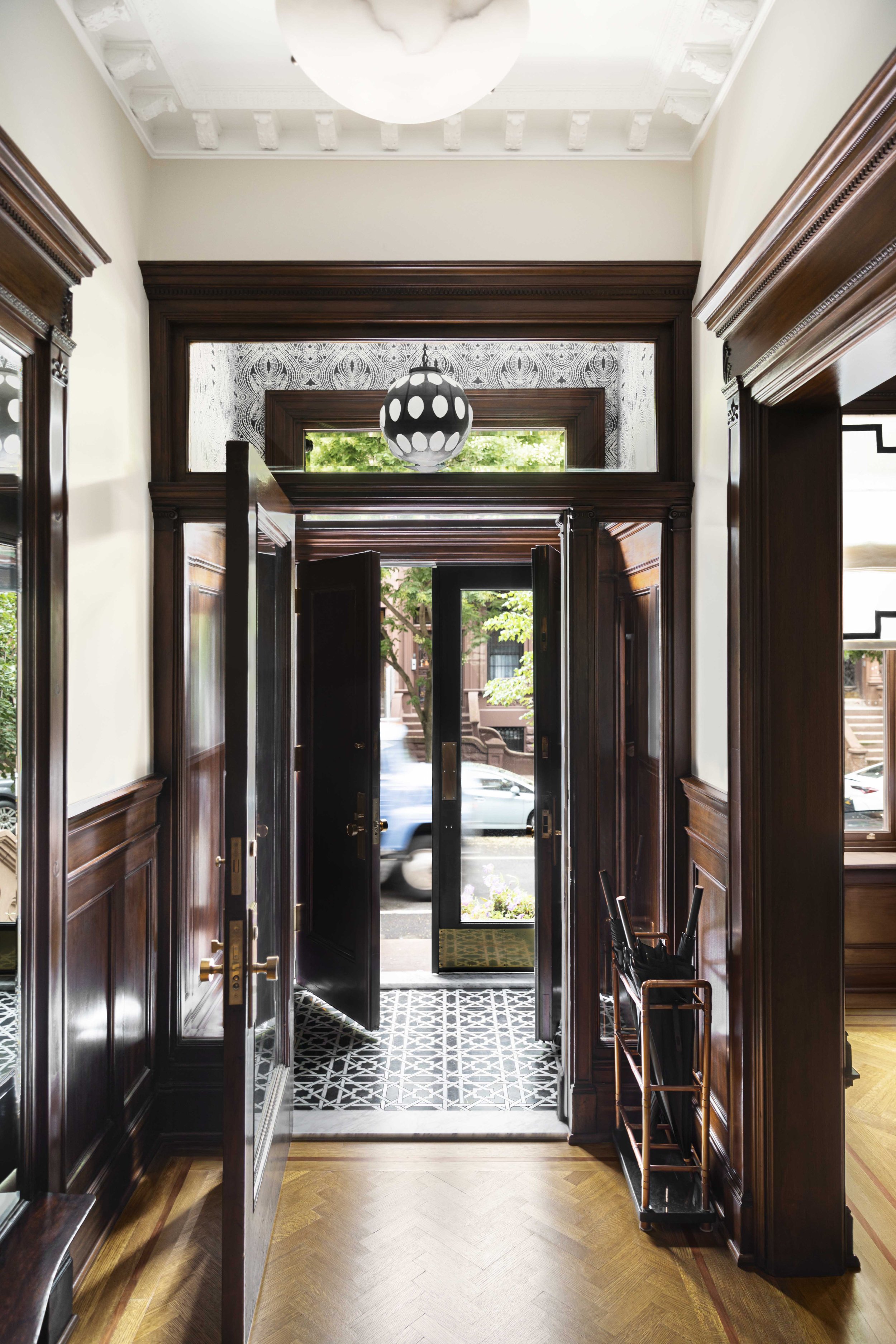 View of a front entrance door with dark wood frame and glass panel, leading to a small porch with trees outside, interior wood paneled walls, high ceilings with intricate molding, and patterned black and white tile flooring at the door.