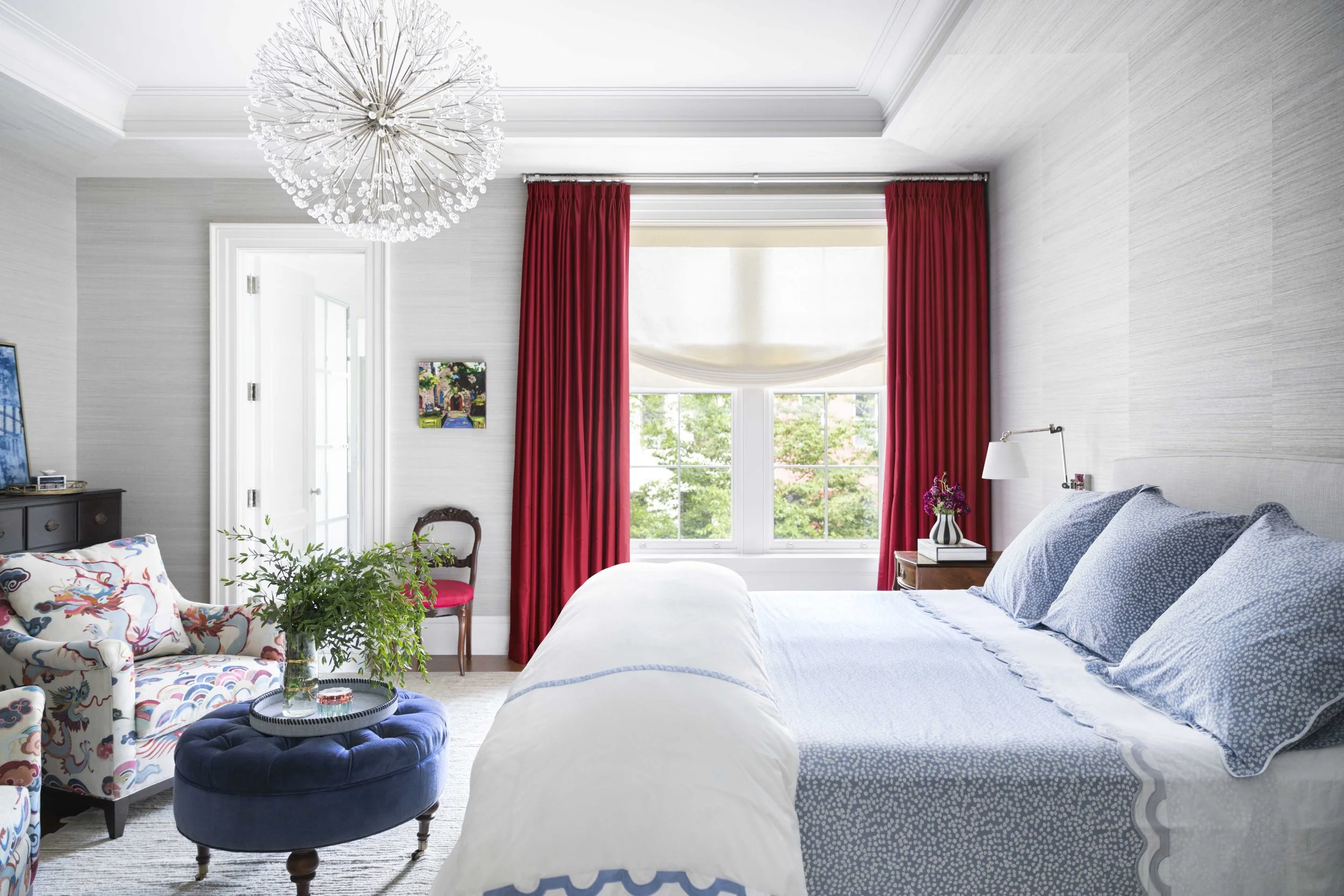 Bright bedroom with white walls, red curtains, a large window with a view of green trees, a bed with blue and white bedding, a pink and white armchair, a dark wood dresser, a small side table with a vase of flowers, and a modern chandelier.