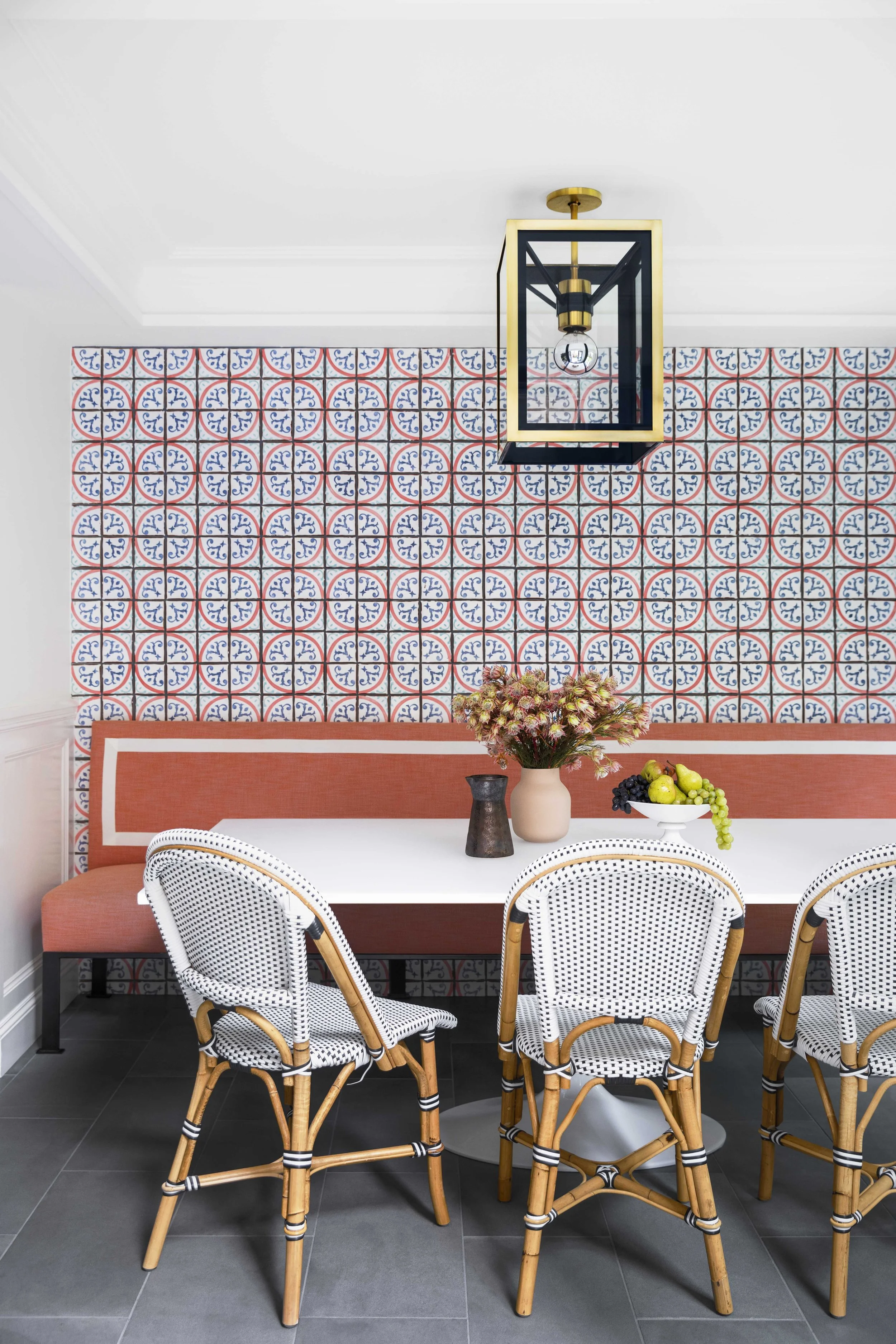 A dining area with a white table, four woven rattan chairs, a built-in bench with pink upholstery, decorative vases, a bowl of fruit, and a patterned tile accent wall with a hanging geometric chandelier.