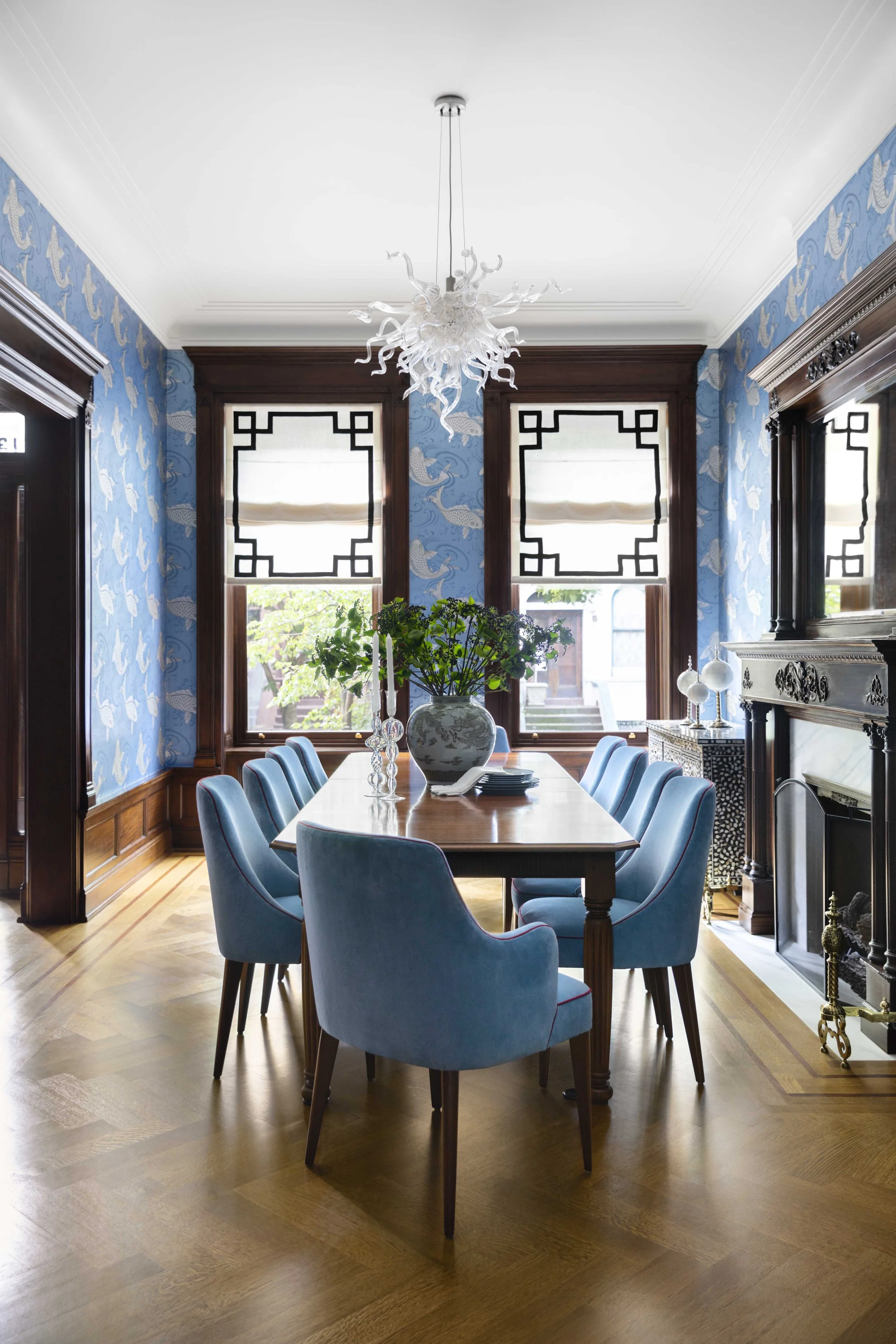 A formal dining room with a wooden table, blue upholstered chairs, a large floral centerpiece, and patterned blue wallpaper. There is a decorative chandelier hanging from the ceiling and a fireplace on the right side.