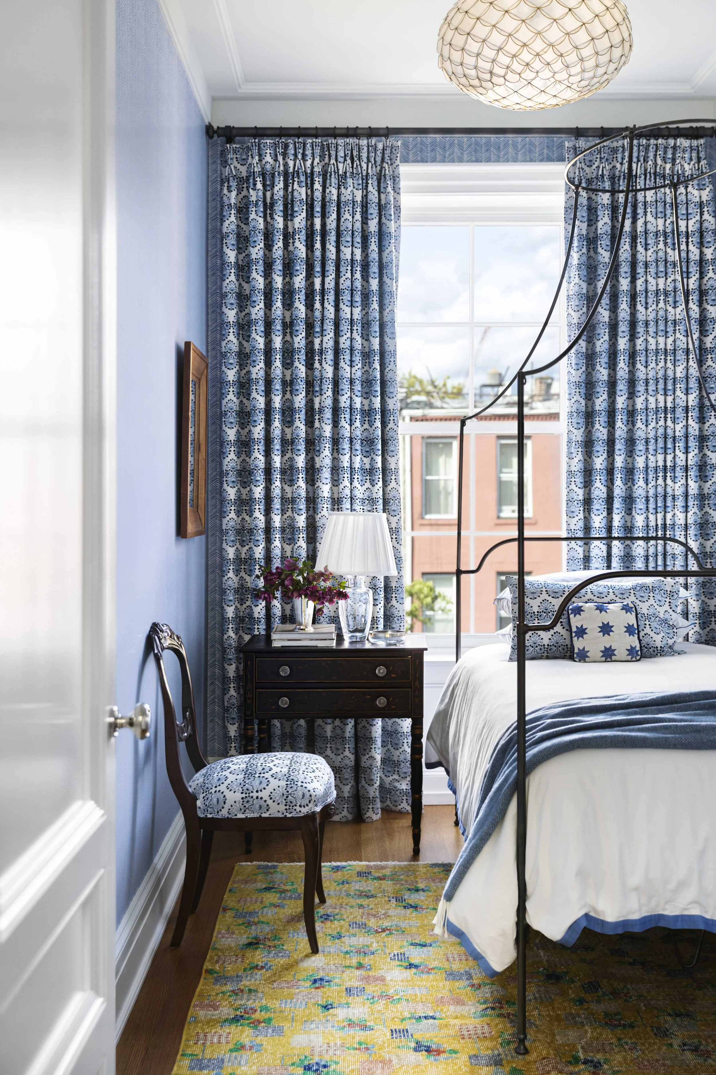 A bedroom with blue patterned curtains, blue and white bedding, a wooden side table with a lamp and flowers, a chair with a matching fabric, a large window showing a cityscape, and a decorative ceiling light.