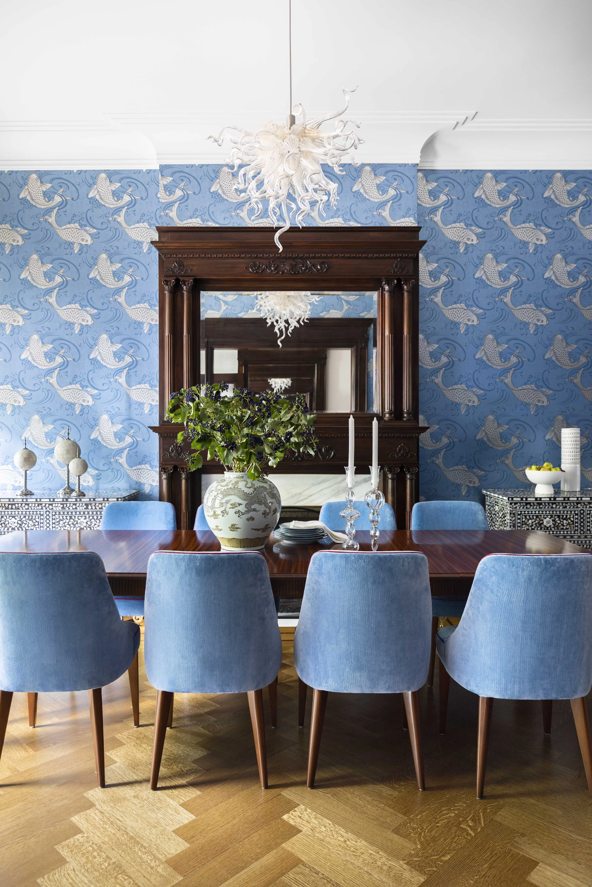 A dining room with a wooden table surrounded by six blue upholstered chairs, a large mirror with a dark wooden frame on a blue wallpapered wall, a white chandelier hanging from the ceiling, and decorative items including a large vase with green leave