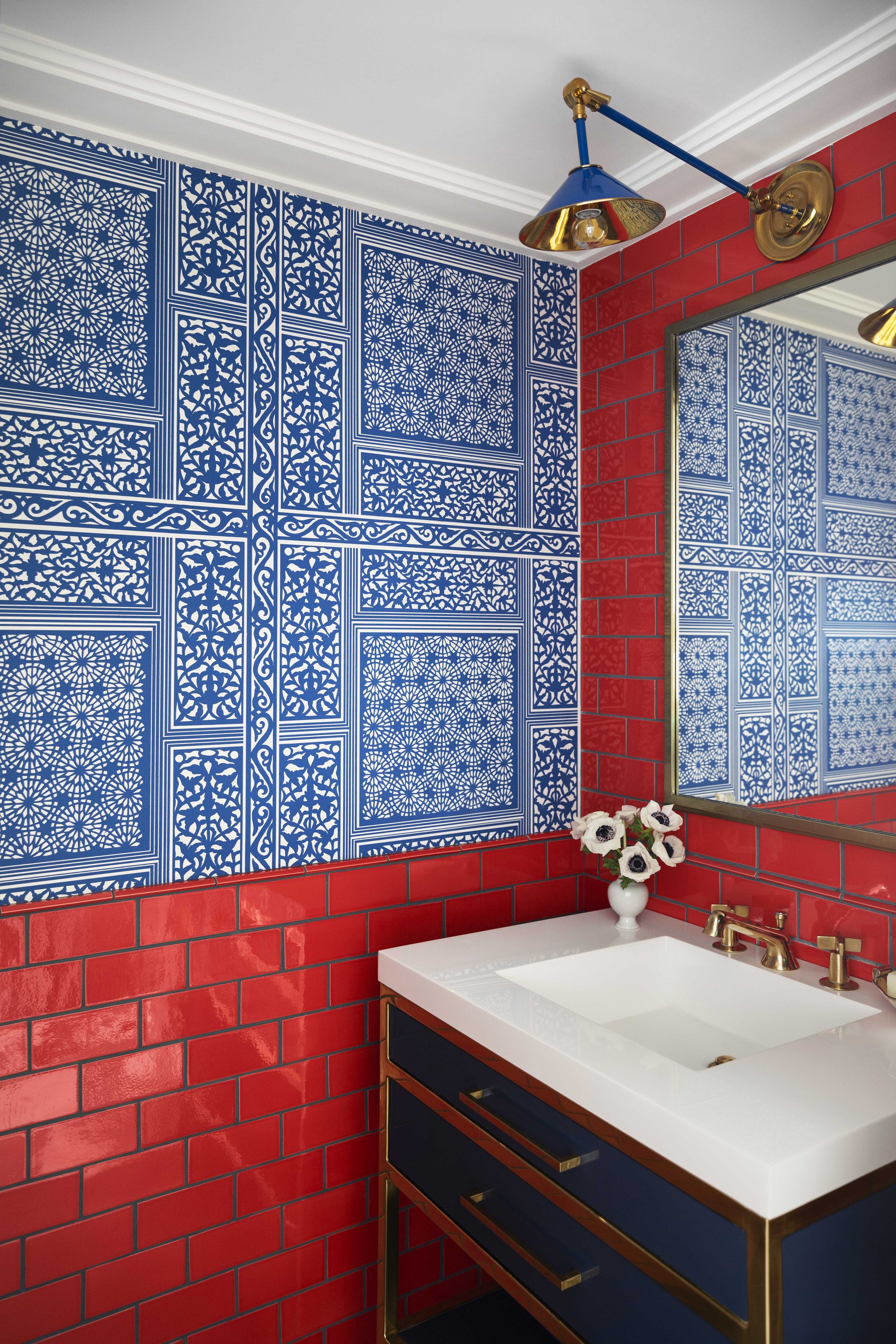 Bathroom with red tiled walls, a boat-shaped white sink, a navy blue vanity with gold handles, a glass-fronted mirror, a blue and gold wall lamp, and a vase with white flowers on the sink.