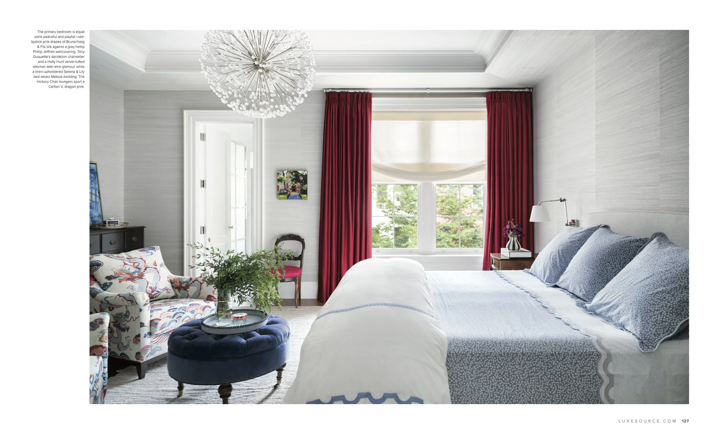 Bright bedroom with a large window, red curtains, and a bed with blue patterned bedding. Decor includes a floral armchair, a dark dresser with a TV, a small side table with flowers, and a contemporary chandelier.