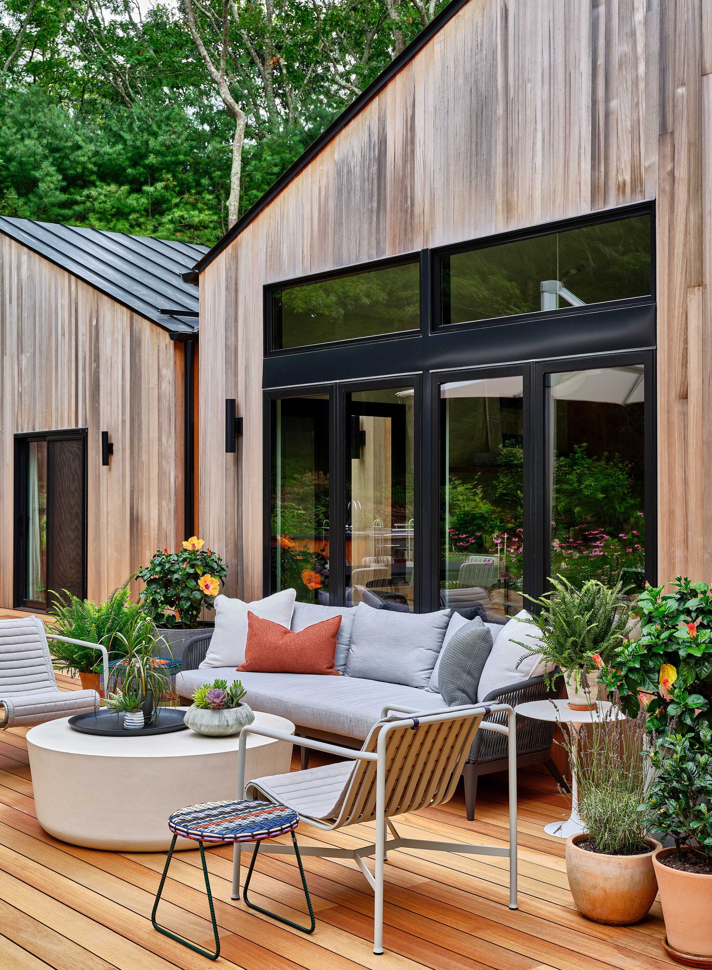 Outdoor patio with wooden decking, a modern sofa with colorful cushions, and several potted plants, in front of a contemporary house with large glass windows and wooden exterior.