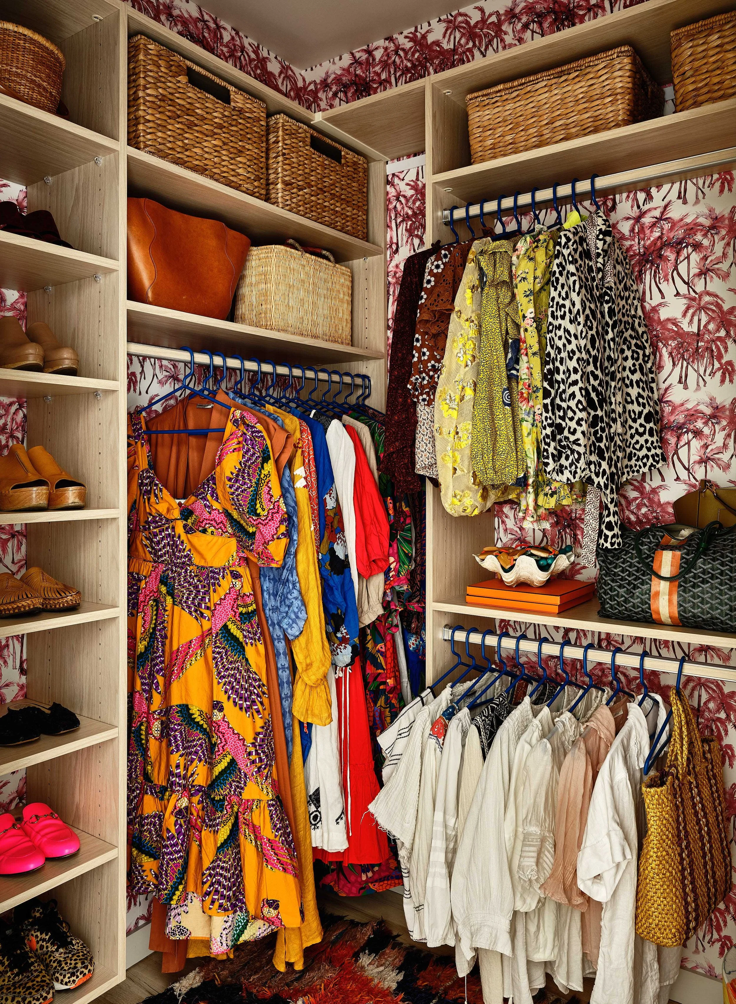 Closet with shelves of woven baskets, a rack of colorful dresses, a rack of white and pastel shirts, and various shoes and accessories, with tropical palm wallpaper.