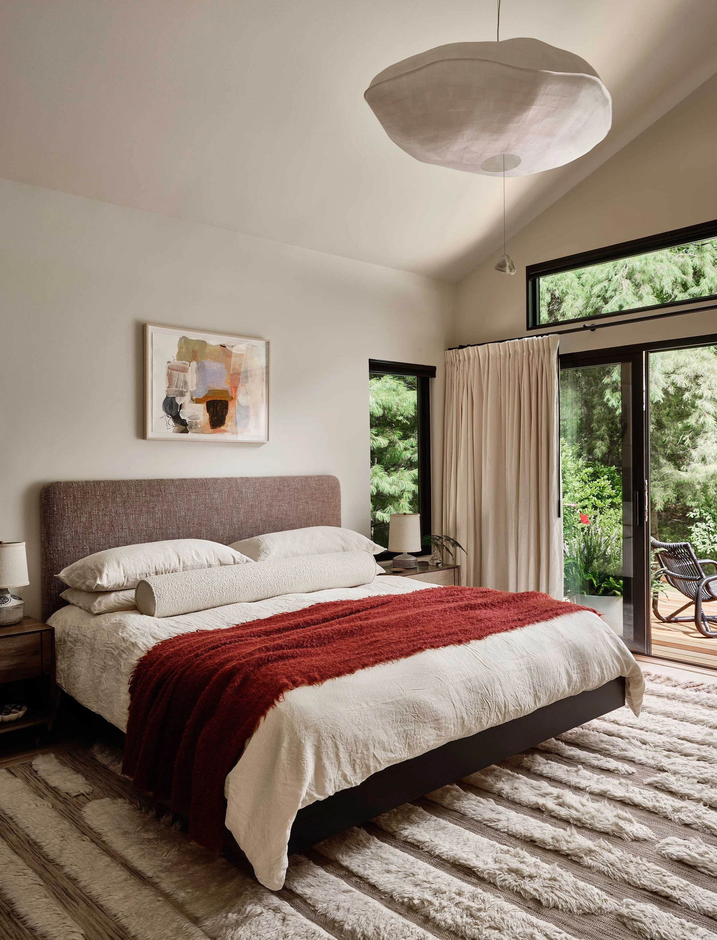 Modern bedroom with a large bed, beige bedding, rust-colored throw, and a textured headboard, next to a window with white curtains overlooking a lush green garden.