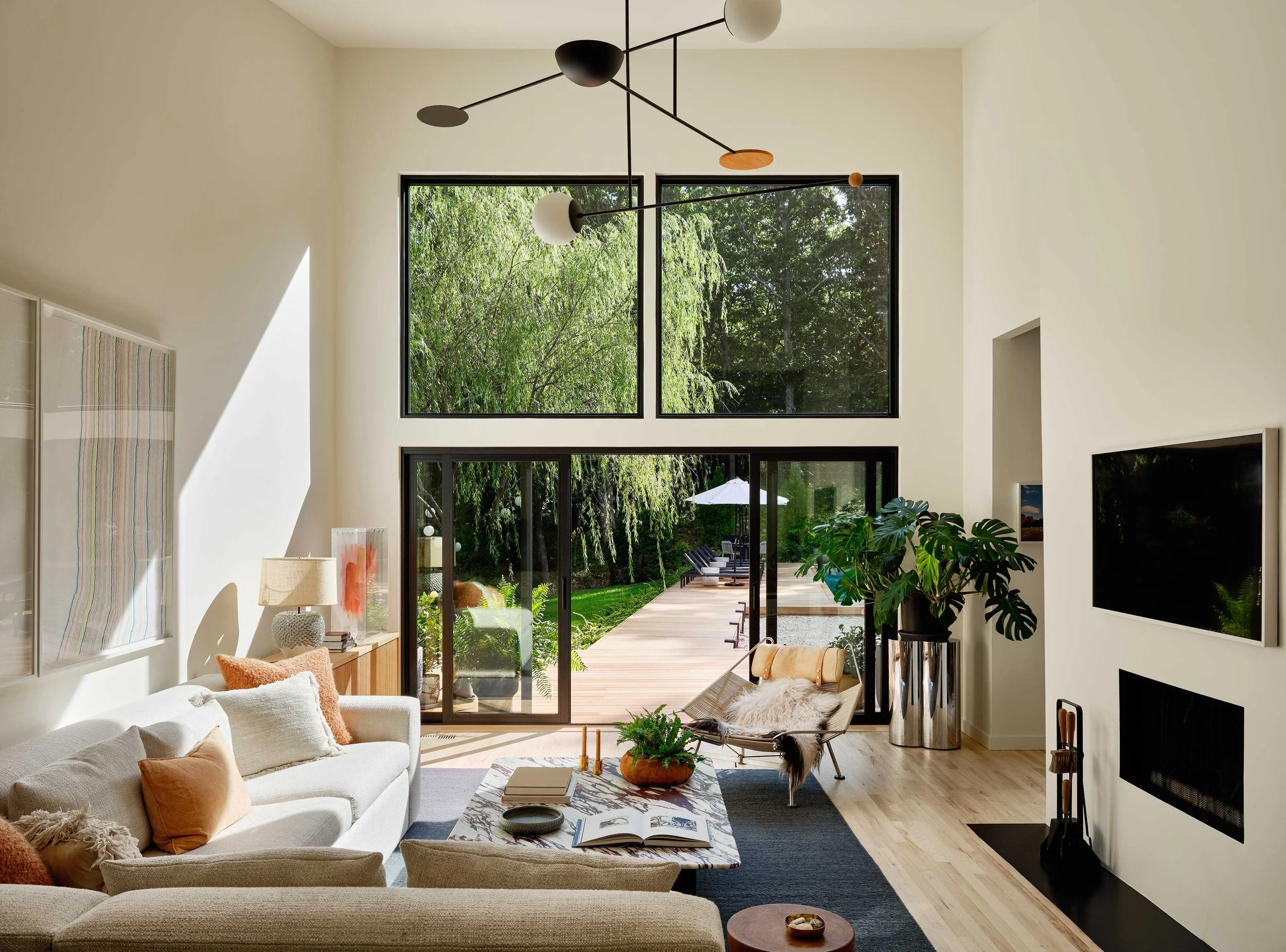 Modern living room with large windows and sliding glass doors overlooking a lush green outdoor patio; white sofas with orange pillows, a marble coffee table with books and a plant, a cream chair with a blanket, and a tall potted plant; artwork on the