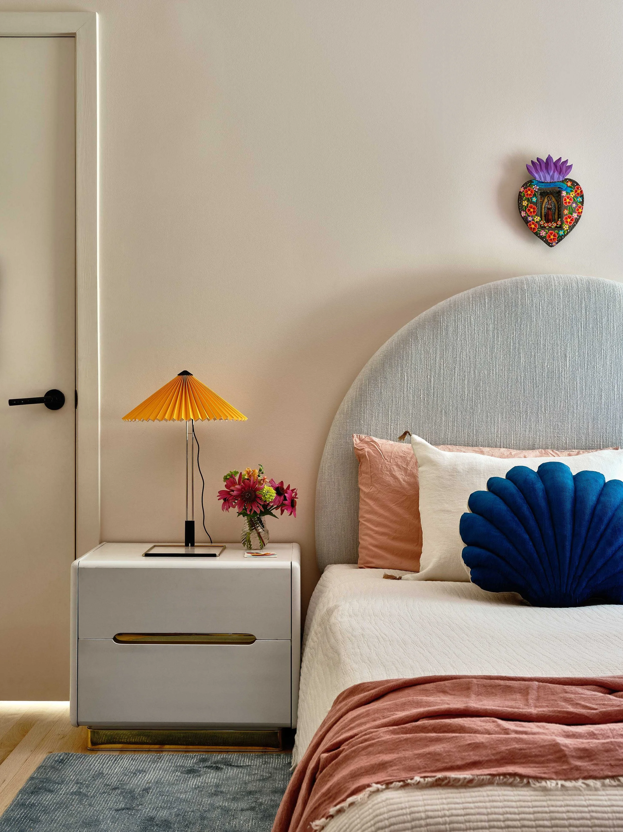 A bedroom featuring a white bed with a light pink blanket and a navy blue shell-shaped pillow. Next to the bed is a white nightstand with a yellow pleated lampshade table lamp and a small vase with pink flowers. The wall behind has a decorative heart