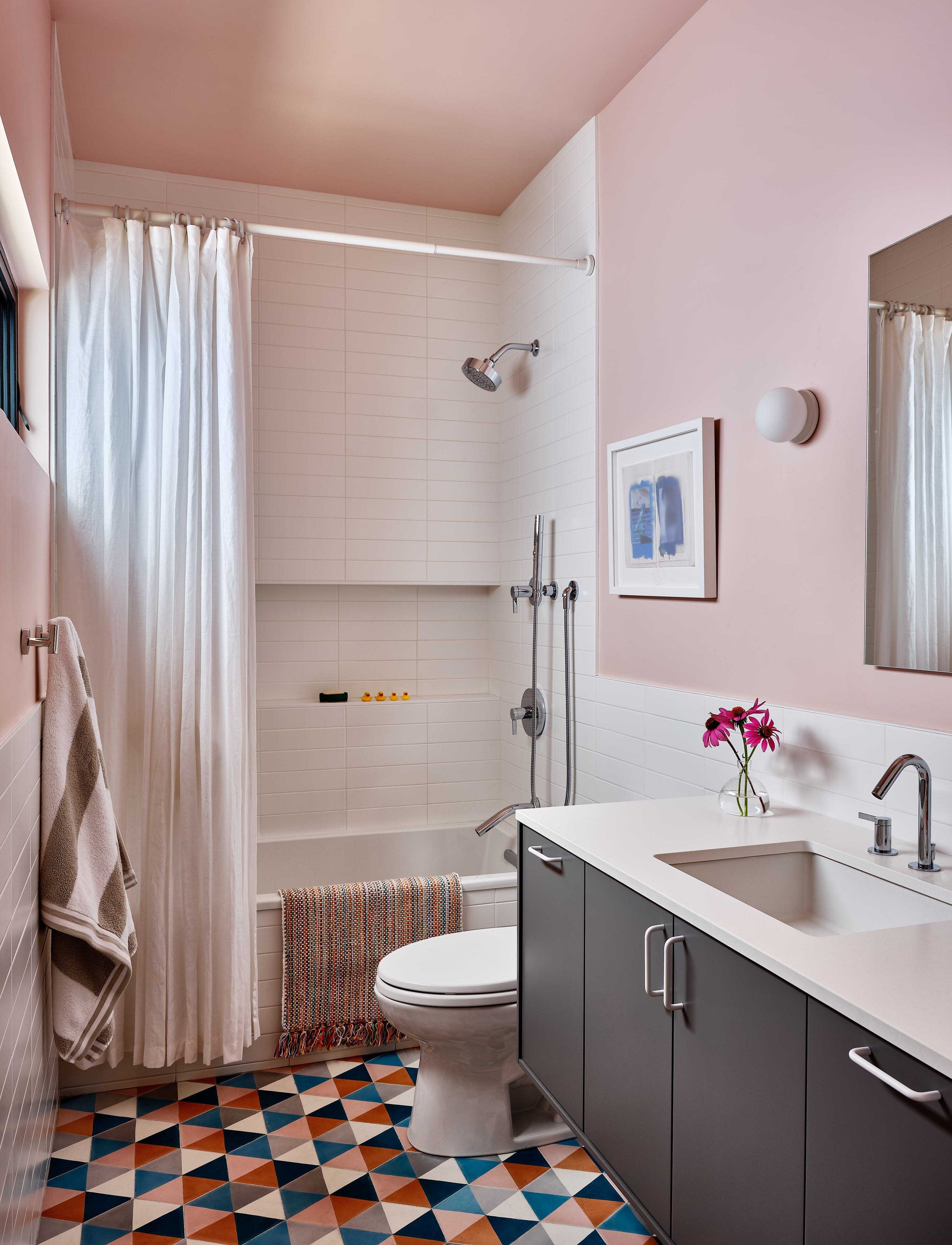 Bathroom with pink walls, shower area, white tile, dark gray vanity, small mirror, colorful patterned floor, and a towel hanging on the wall.