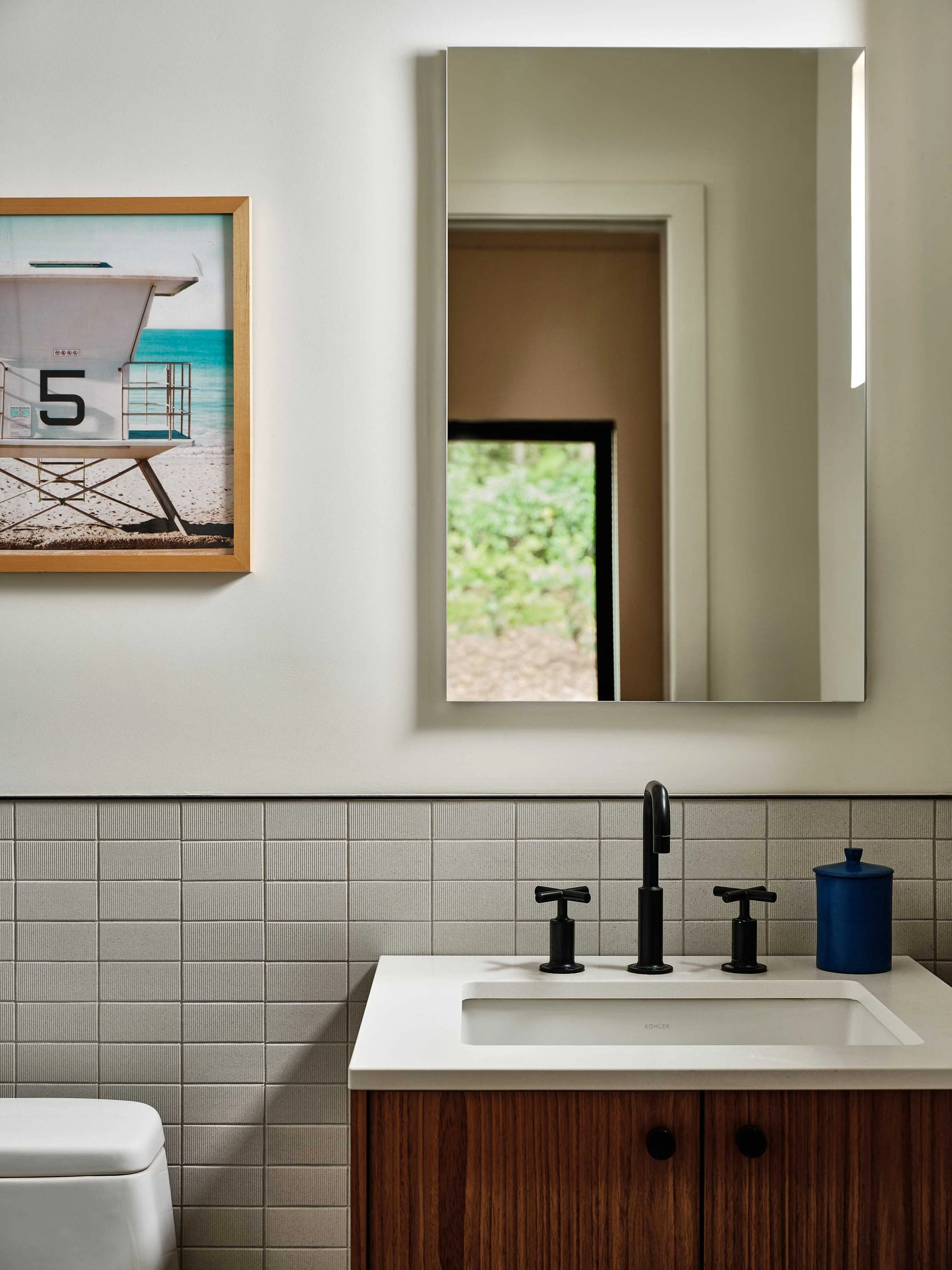 A modern bathroom with a white wall, beige tiled lower wall, a pendant mirror, black faucet, white sink with a wooden cabinet, a blue container, and a framed beach photo on the wall.