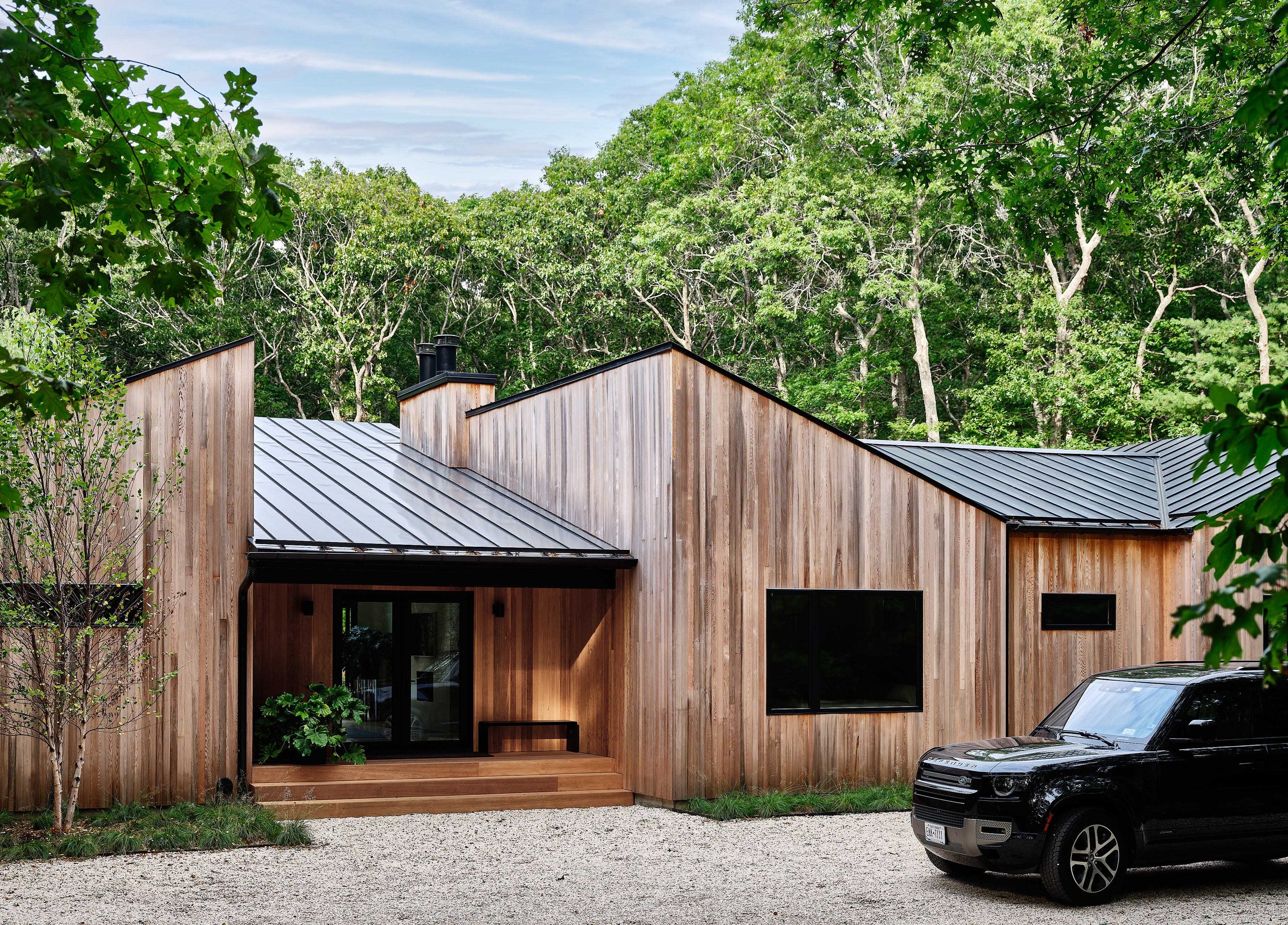 Modern house with vertical wooden siding, black metal roof, large front window, and green landscaping, surrounded by trees, with a black vehicle parked in the gravel driveway.