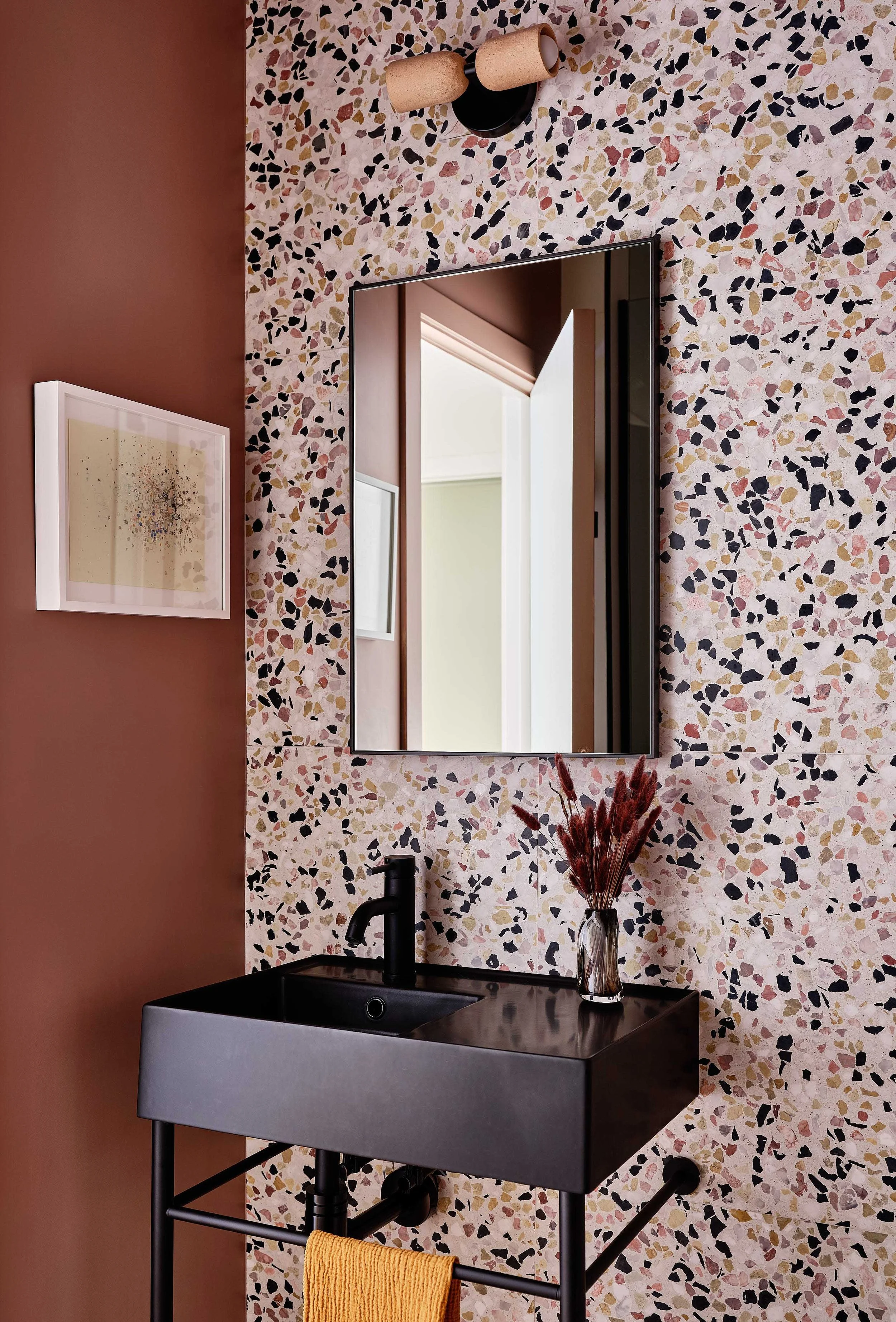 A modern bathroom with a terrazzo wall, a rectangular black sink, a mirror, wall-mounted light fixture, a wall art piece, and a vase with dried red plants on the sink.