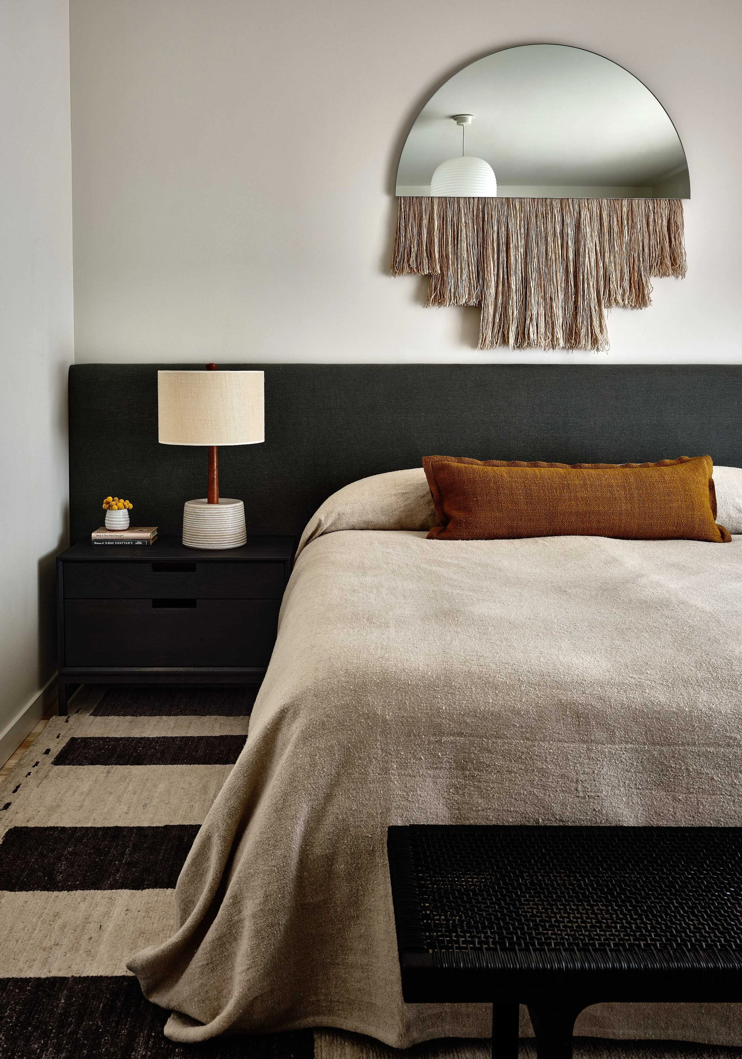 A bedroom with a beige bedspread, rust-colored pillow, black nightstand with a table lamp, small vase with yellow flowers, and books. A half-moon mirror with a woven fringe is on the wall above the bed.
