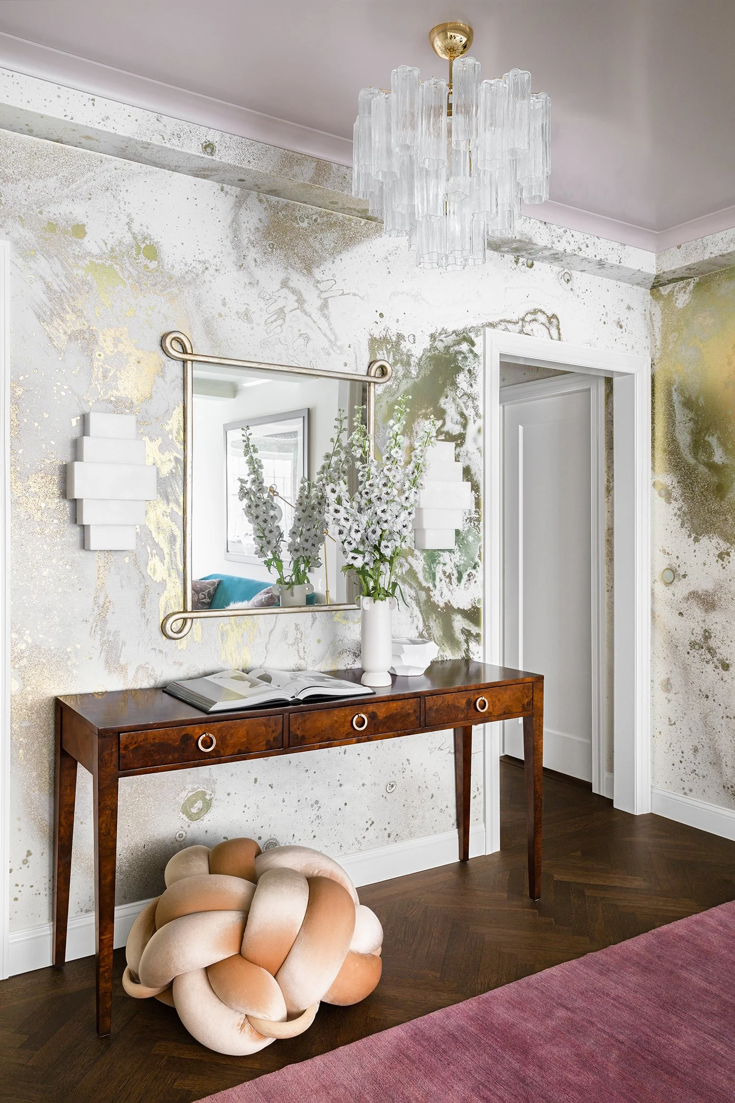 Interior hallway with a wooden console table, mirror, decorative white flowers, and abstract wall decor, hardwood floor, and a chandelier.
