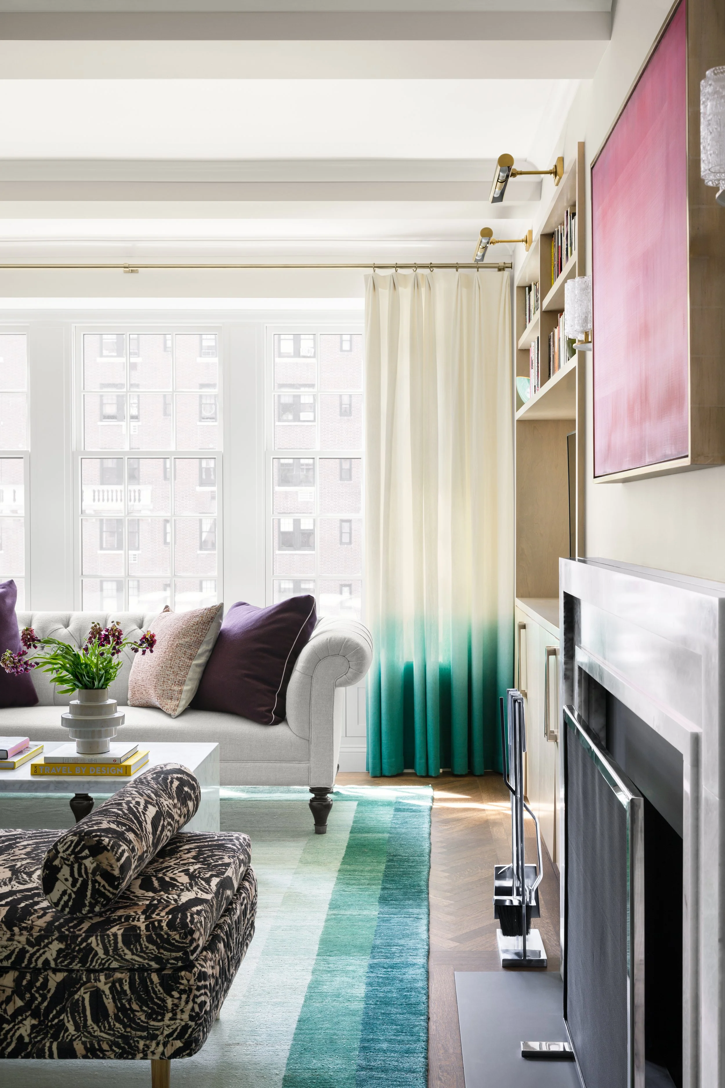 Bright living room with large windows, white curtains, a beige sofa with purple and beige pillows, a patterned armchair, a coffee table with a plant and books, a colorful rug, a fireplace, and built-in shelves with books.