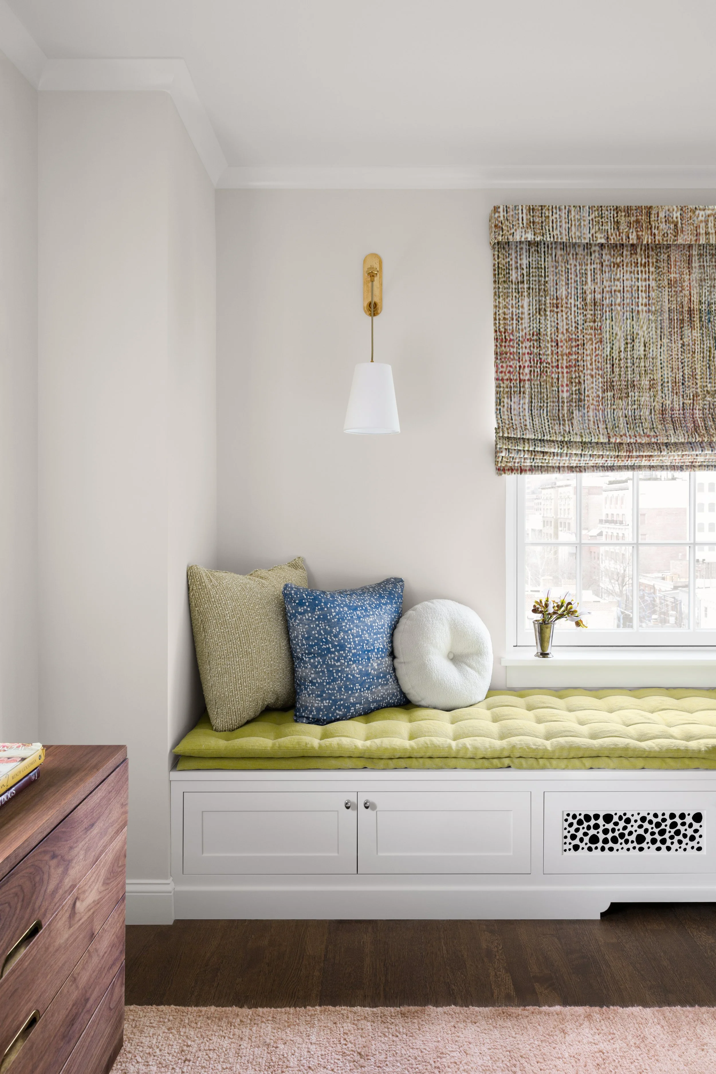 A cozy window seat with a yellow tufted cushion, three pillows, a small potted plant, and a window with patterned Roman shade in a bright room.