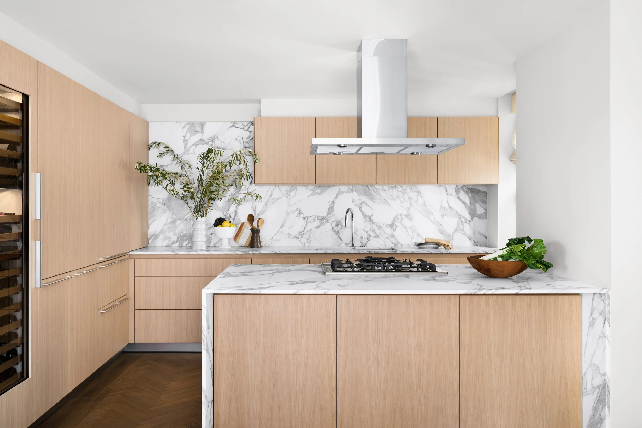 Modern kitchen with light wood cabinets, marble countertops, and greenery.