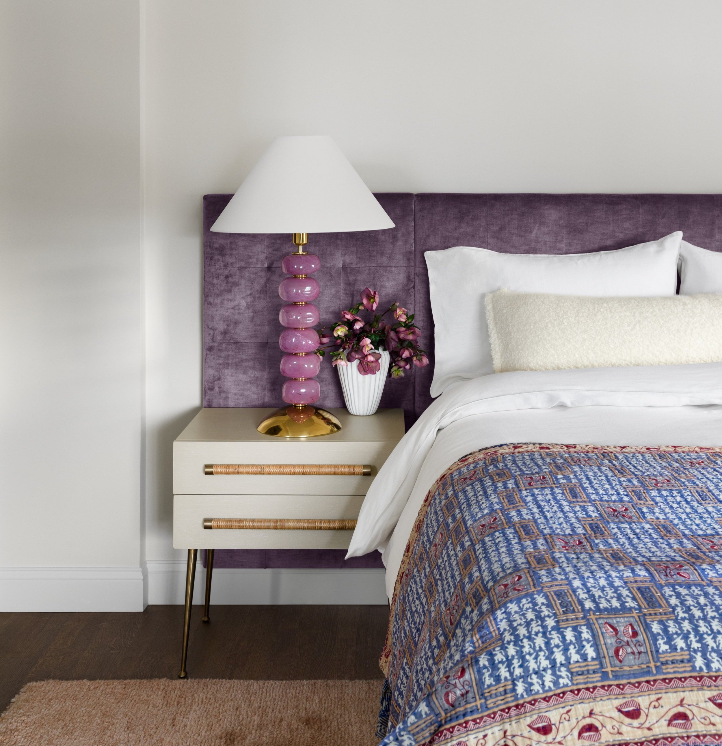 A bedroom corner with a purple velvet headboard, a nightstand with a pink glass lamp, a white and pink flower vase, a white pillow, a textured cream pillow, and a bed with blue, red, and beige patterned bedding.