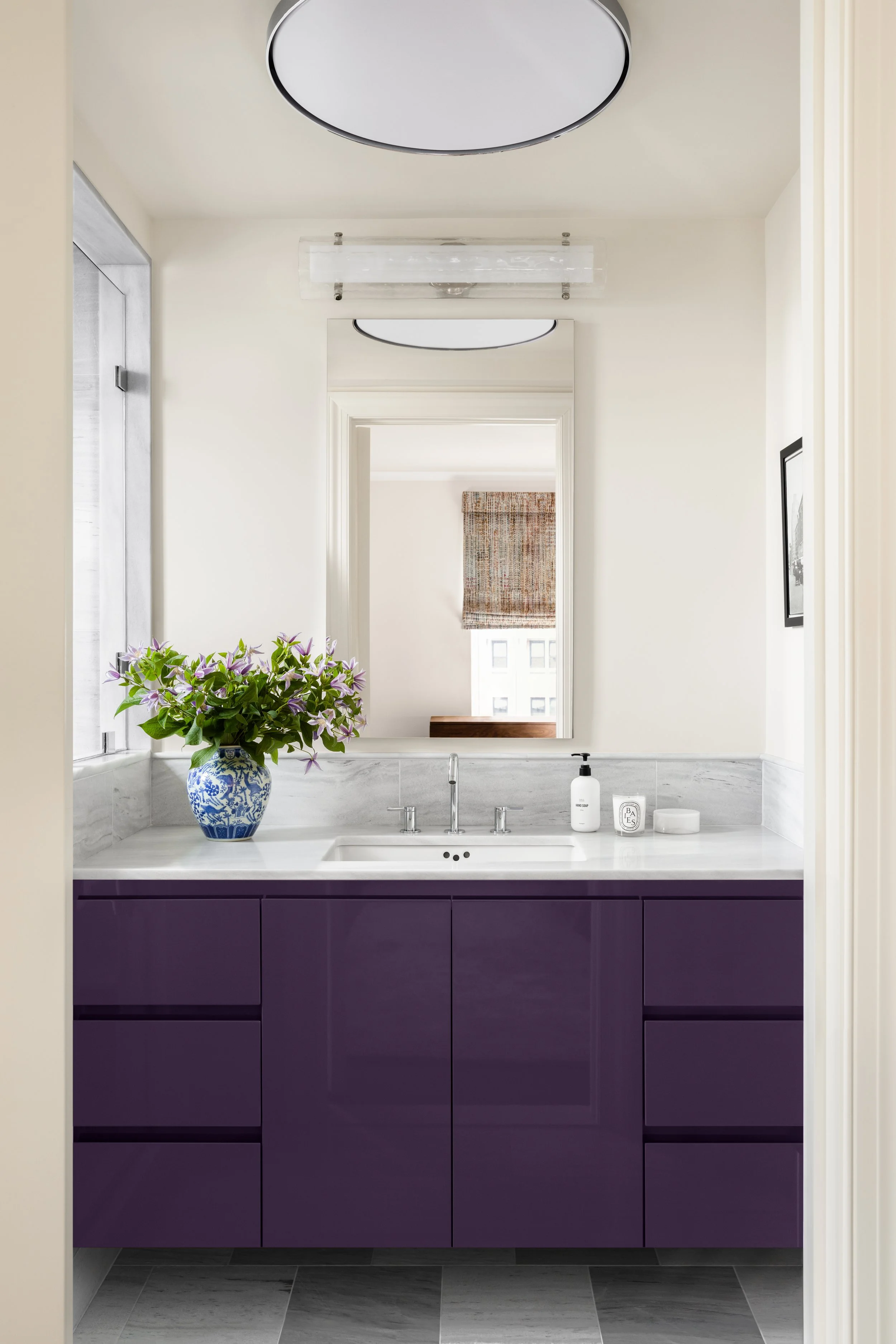 A bathroom with a white marble countertop, purple cabinet, large mirror, and decorative vase with flowers.