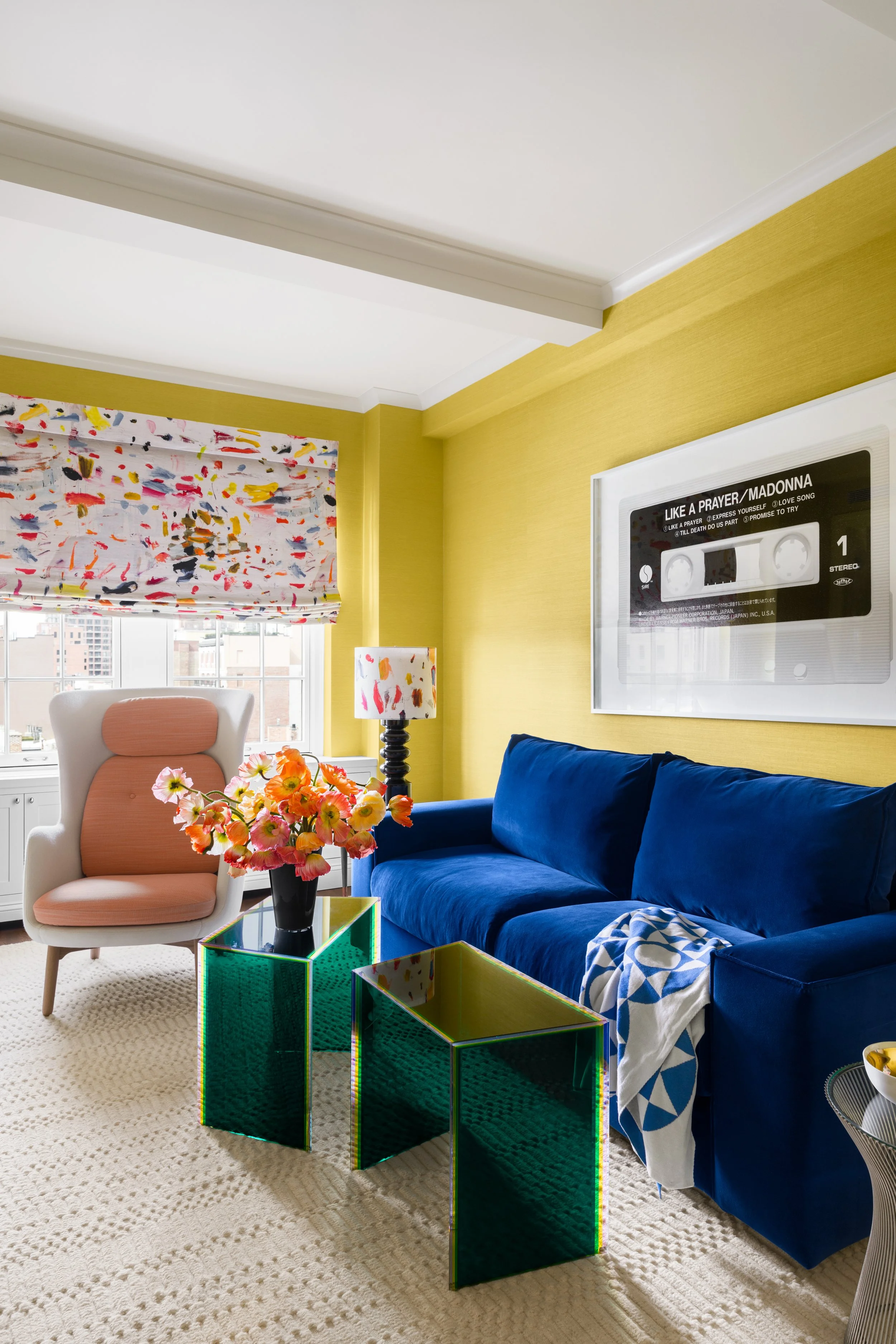 Colorful living room with yellow walls, a large window, a pink and white armchair, a dark blue velvet sofa, a decorative lamp, a bouquet of pink flowers on a green side table, and a wall art piece in the shape of a cassette tape.