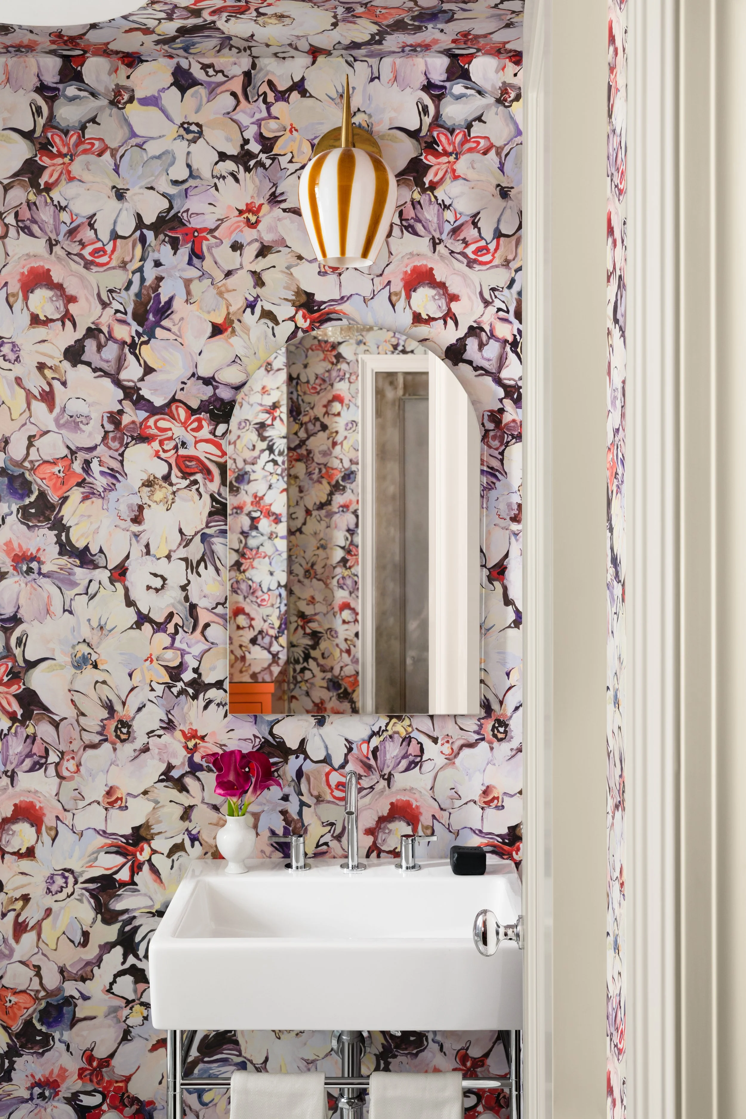 A bathroom with floral patterned wallpaper, a mirror, a white sink with a vase of pink flowers, a wall-mounted soap holder, and a hanging striped gold and white pendant light.