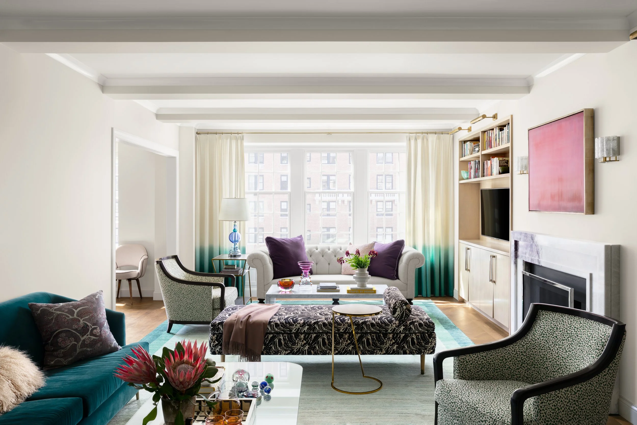Bright living room with beige walls, large windows, white curtains, and decorated with colorful cushions, a fireplace, bookshelves, and modern furniture including a teal sofa, patterned armchairs, and a glass coffee table.