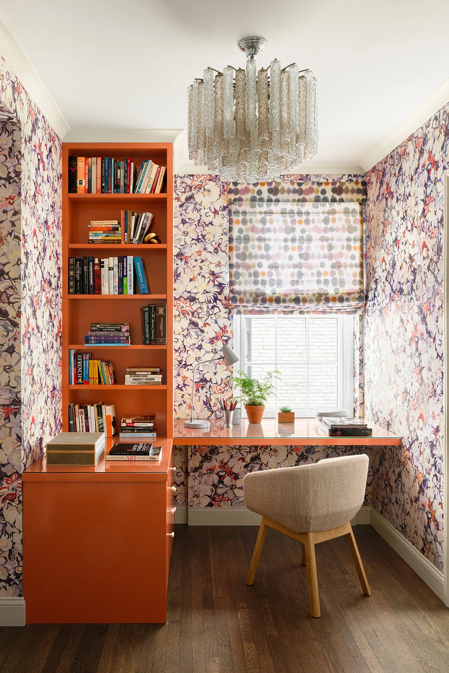 A home office with a coral desk and bookshelf, floral wallpaper, a window with colorful polka-dot roman shade, a beige upholstered chair, decorative items, green plants, and a hanging crystal chandelier.
