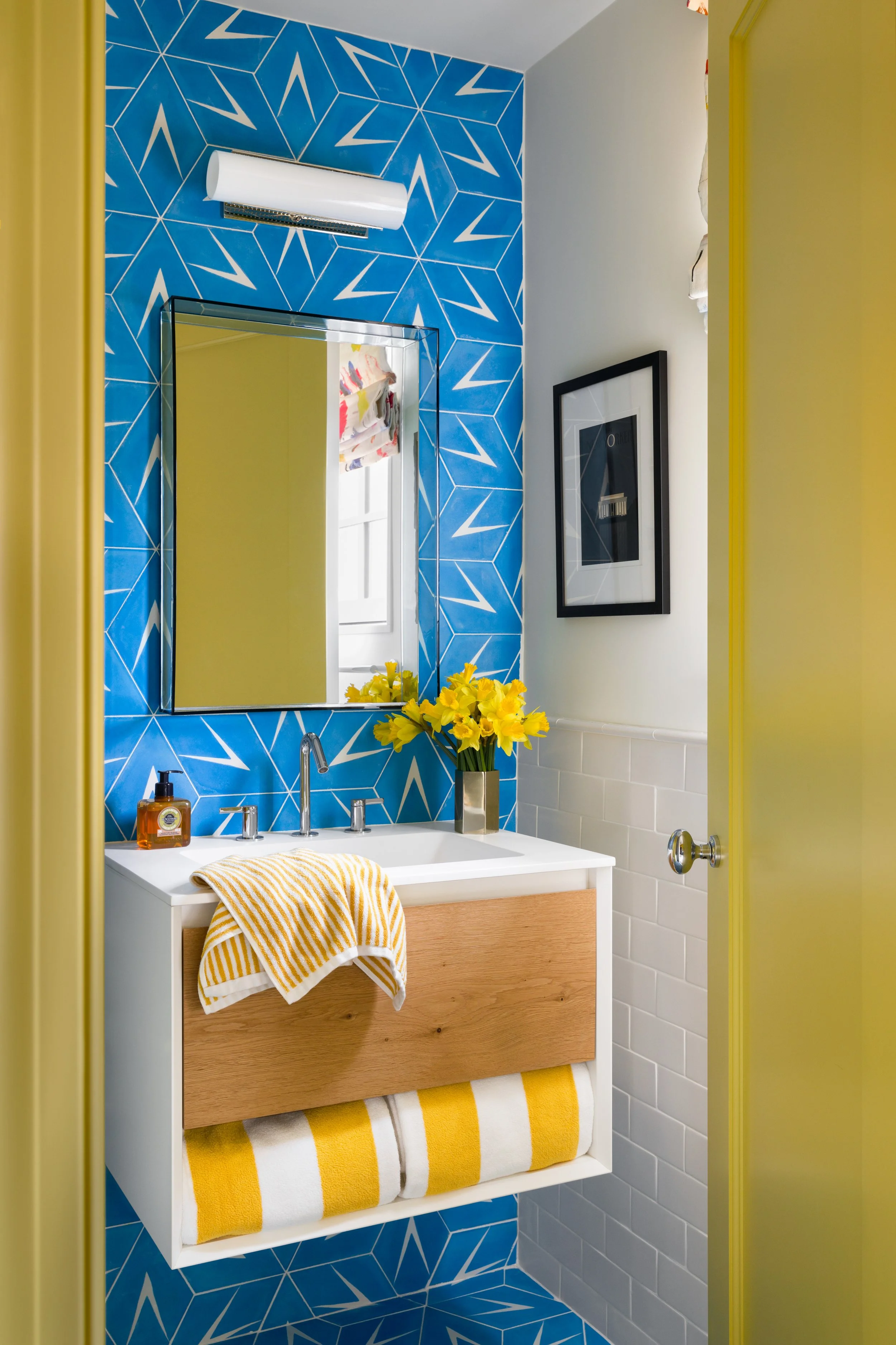 A bathroom with a blue geometric-patterned wall, a mirror, a white vanity with a wooden drawer, a yellow and white striped towel, a soap dispenser, a vase with yellow flowers, and a framed photo on a white wall.