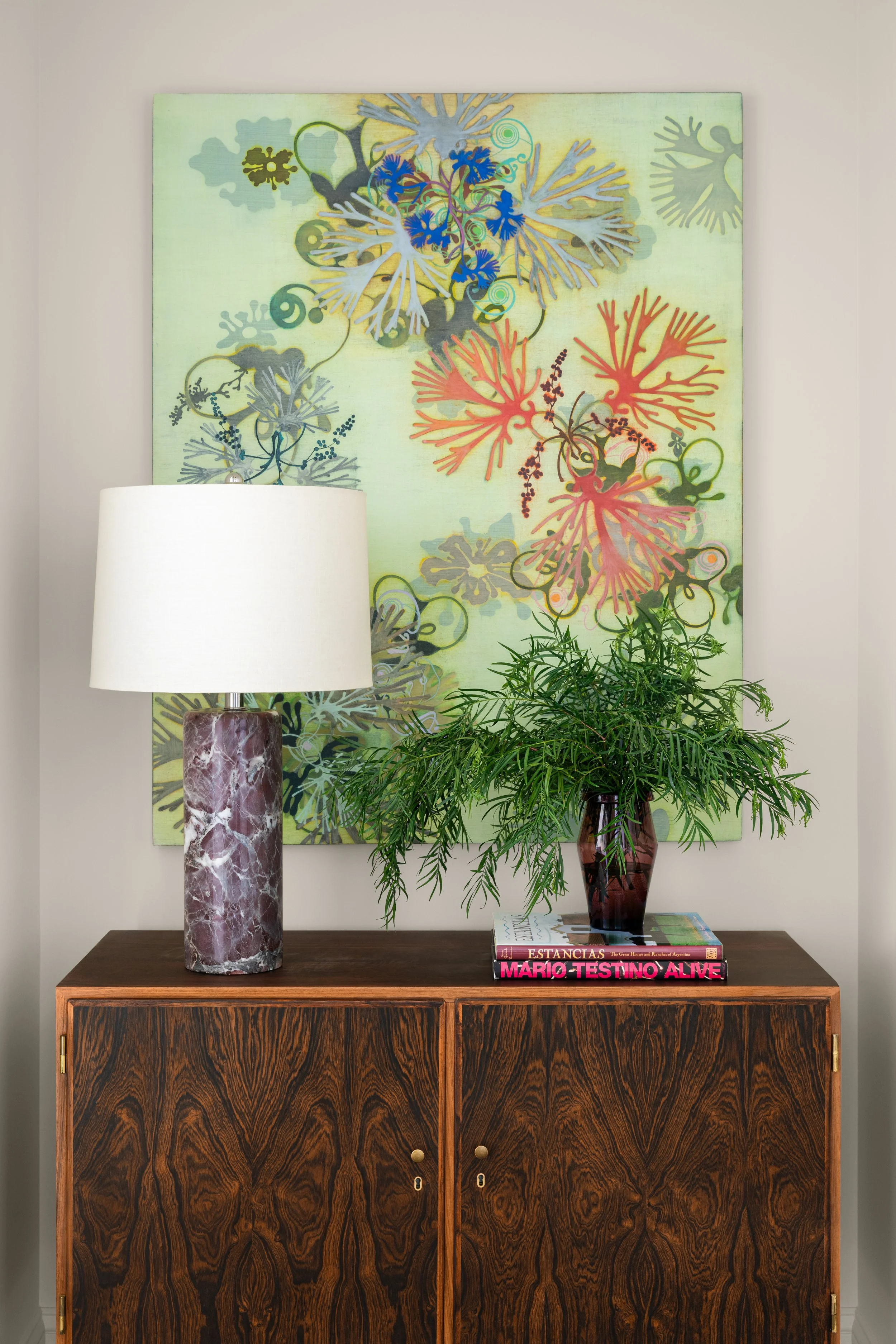 A wooden cabinet with two doors topped with a table lamp with a marbled pink and white base and a white lampshade, a green leafy plant in a dark vase, and a colorful abstract floral painting hanging on the wall behind.