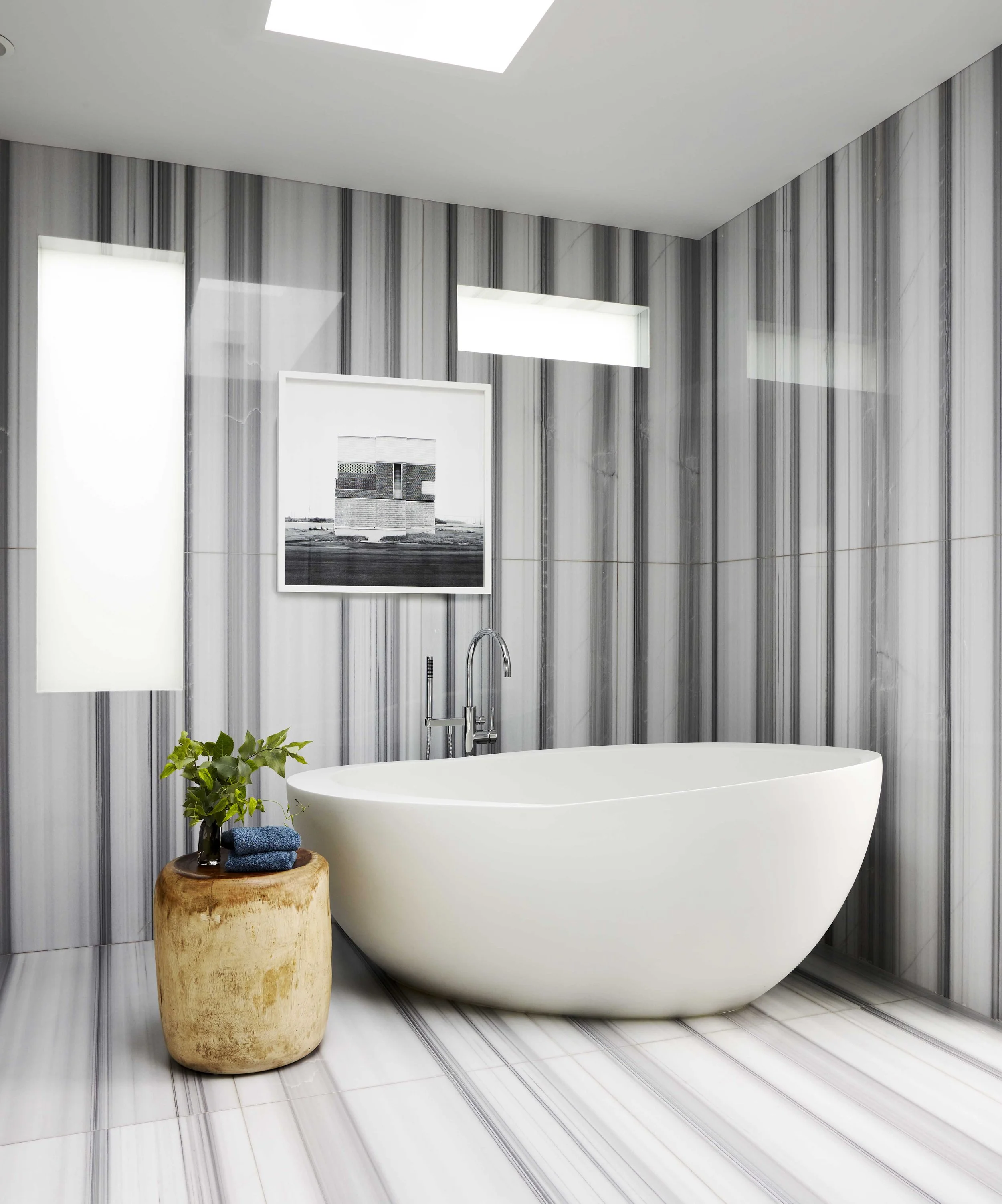 Modern bathroom with a white bathtub, gray striped marble walls, striped marble floors, a small wooden side table with a potted plant and towels, framed artwork on the wall, and an overhead skylight