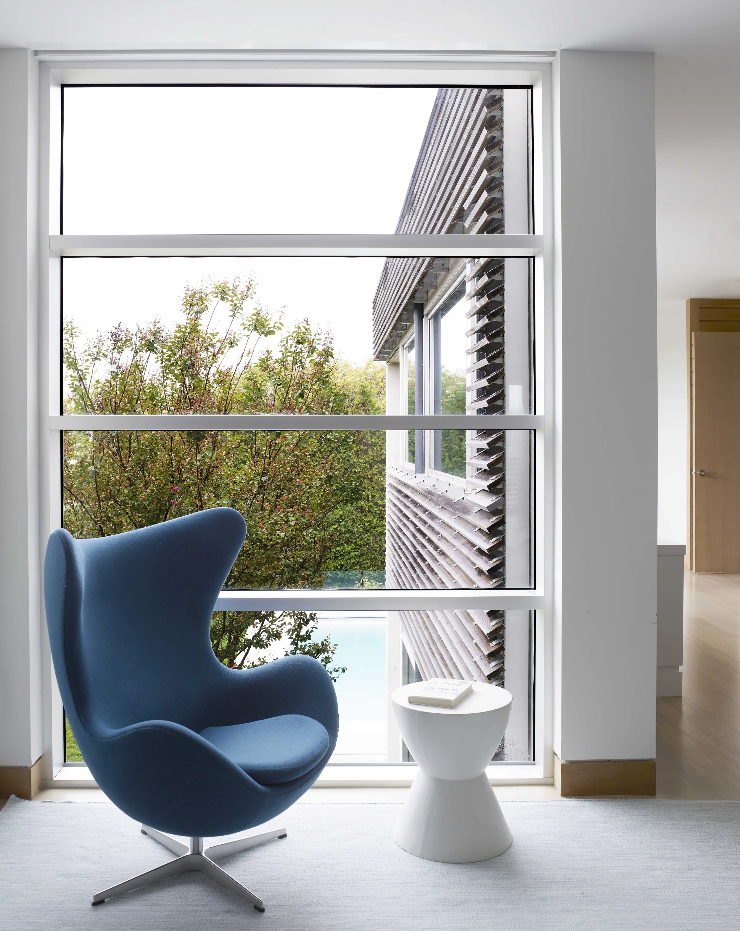 Interior of a modern room with a blue chair, small white side table with a book, large window with view of trees and a neighboring building.