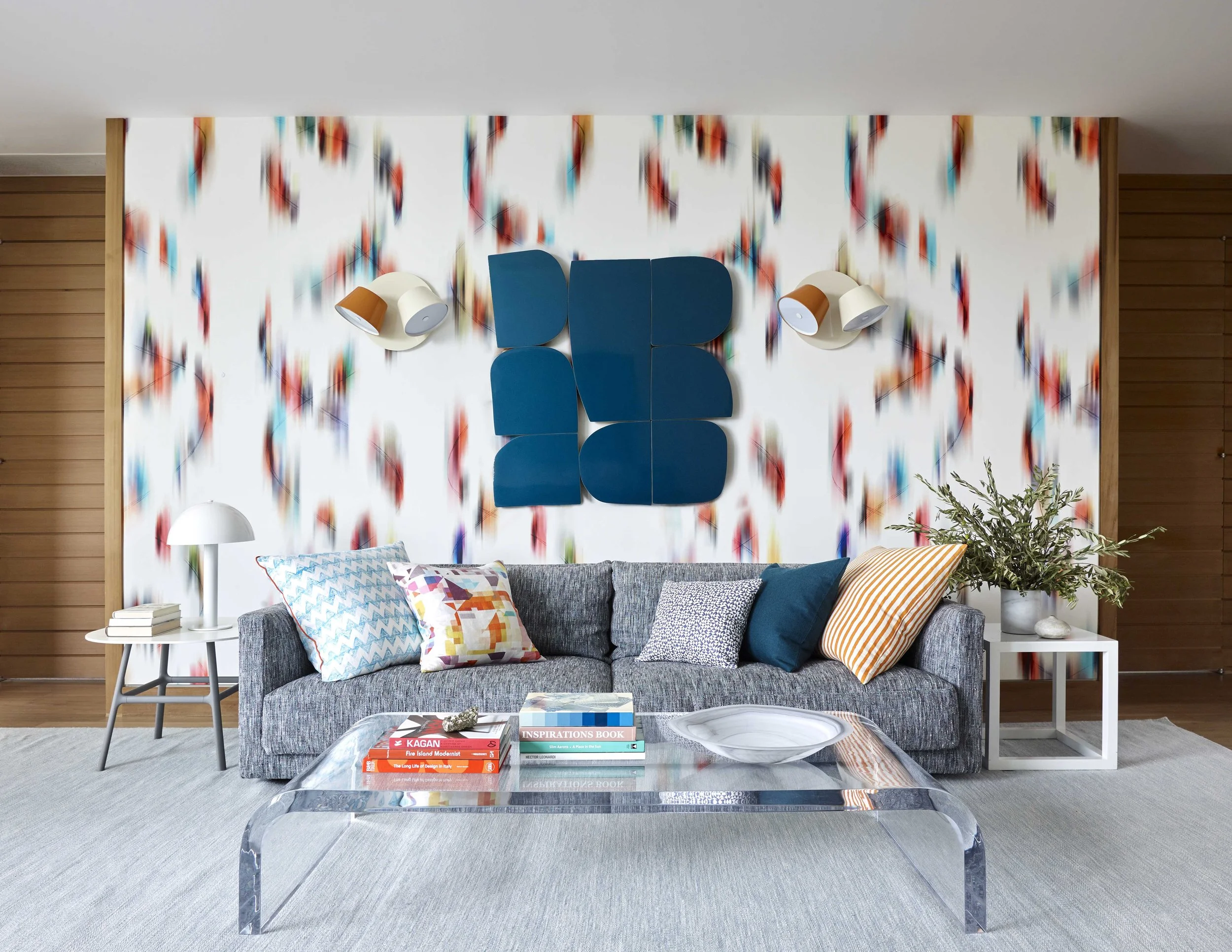 Modern living room with gray sofa, colorful patterned pillows, white side tables, a glass coffee table with books and a tray, arrow-patterned wallpaper, blue wall art, wall sconces, wooden wall paneling, and a large potted plant.