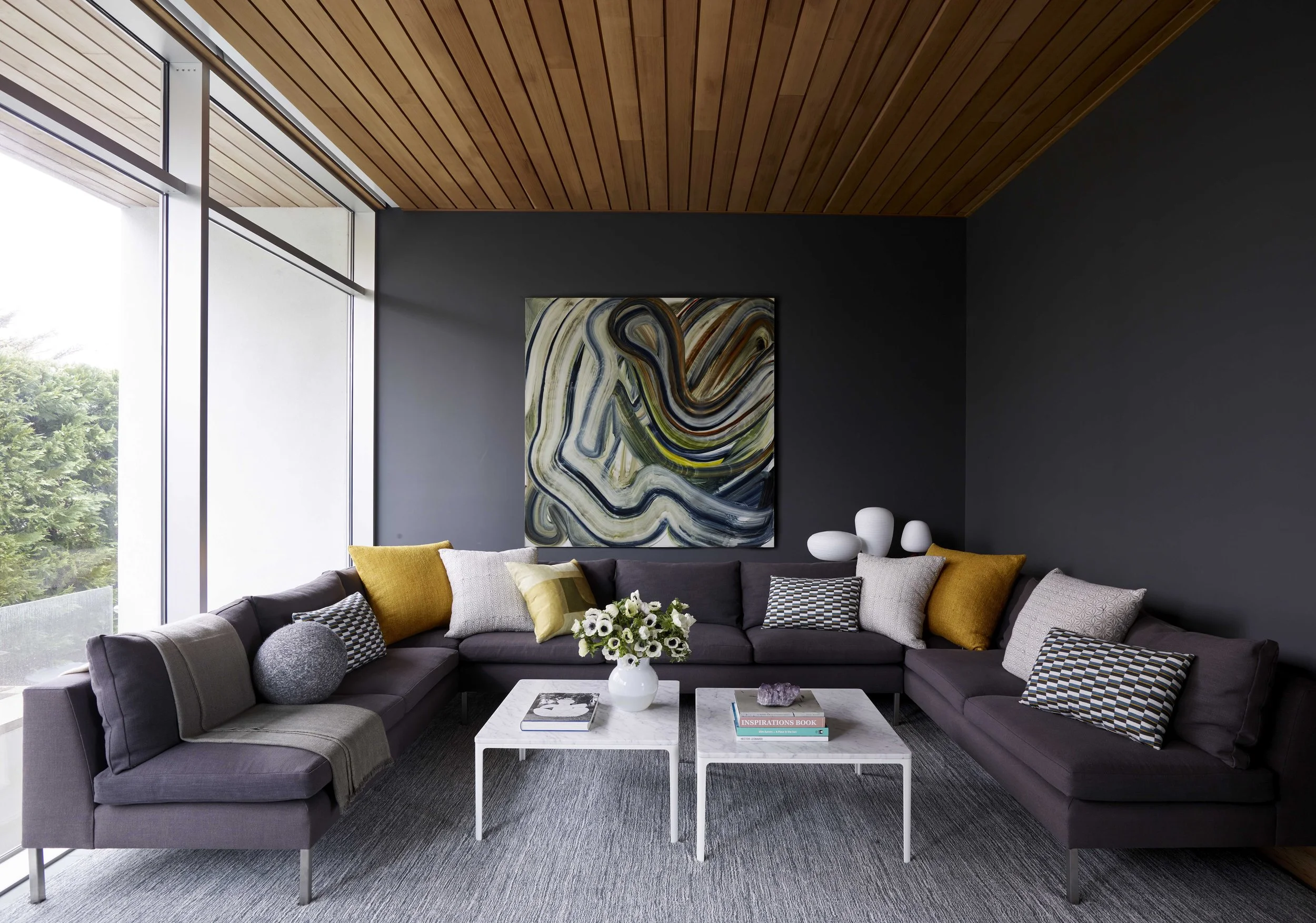 Modern living room with a dark gray sectional sofa decorated with various yellow, beige, and checkered pillows, a white marble coffee table with a vase of white flowers, and abstract art on a dark wall. Large windows bring in natural light.