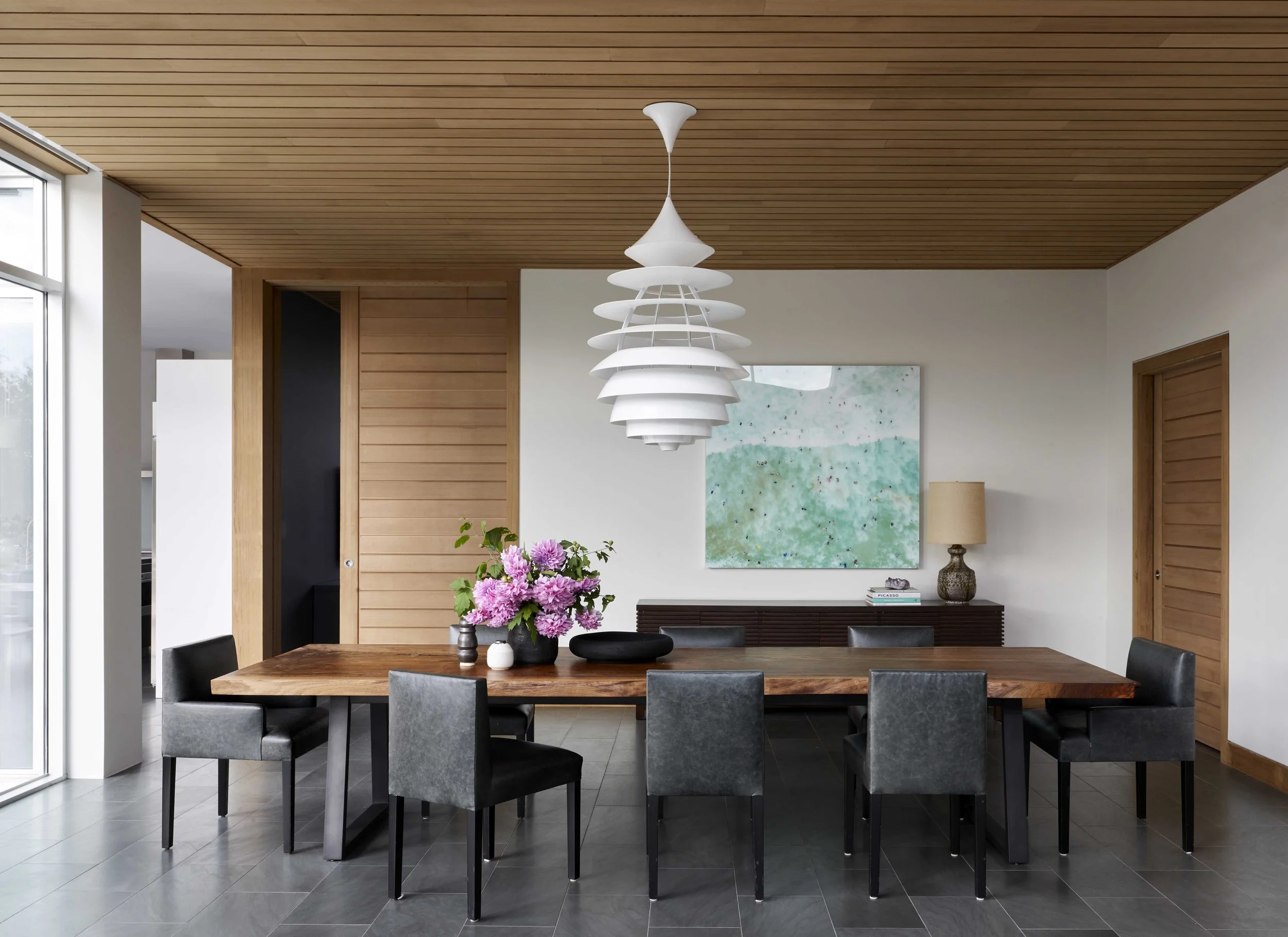 Modern dining room with a large wooden table, eight black and gray chairs, a white layered pendant light, a vase of pink flowers, a sideboard, an abstract painting, a table lamp, and a wood ceiling with natural light from large windows.