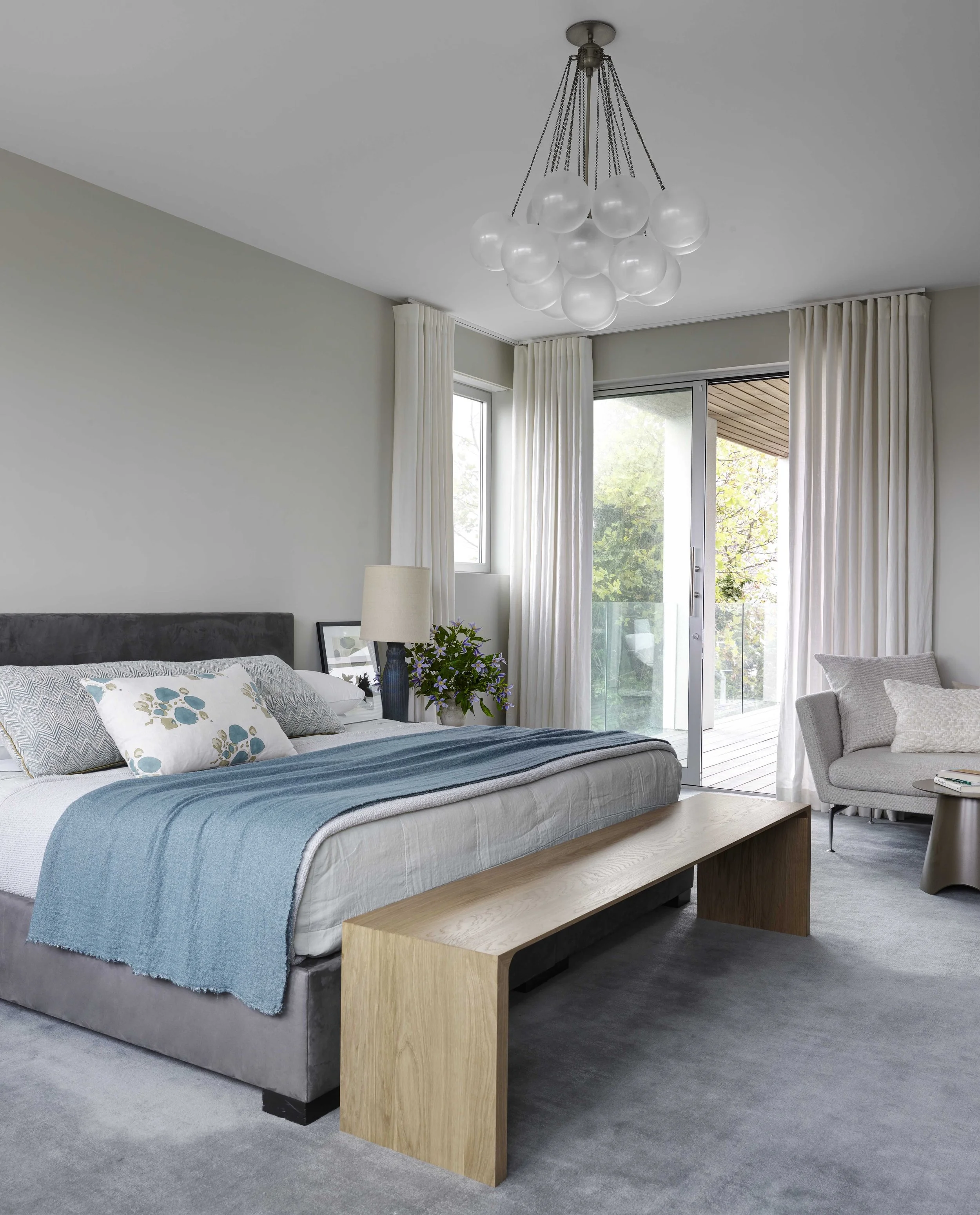 Modern bedroom with bed, wooden bench, armchair, floor-to-ceiling curtains, sliding glass door, ceiling light fixture, and balcony view.