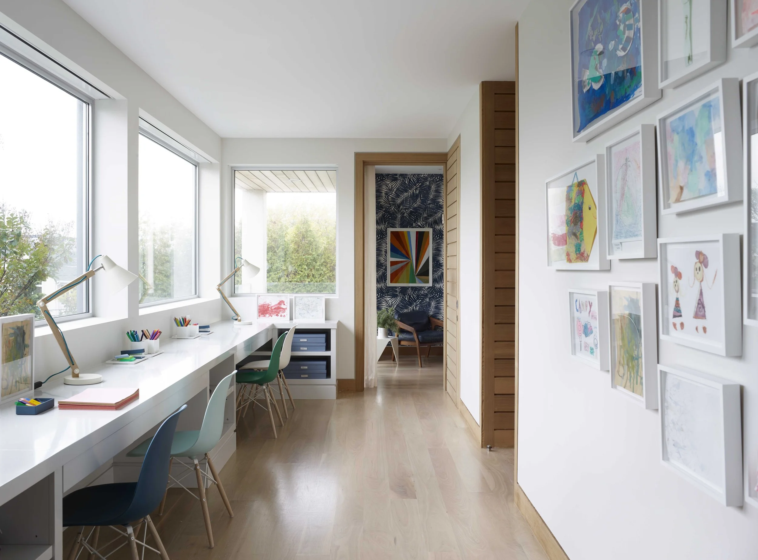 Bright art room with a long white desk along large windows, colorful chairs, desk lamps, and children's artwork on the walls.